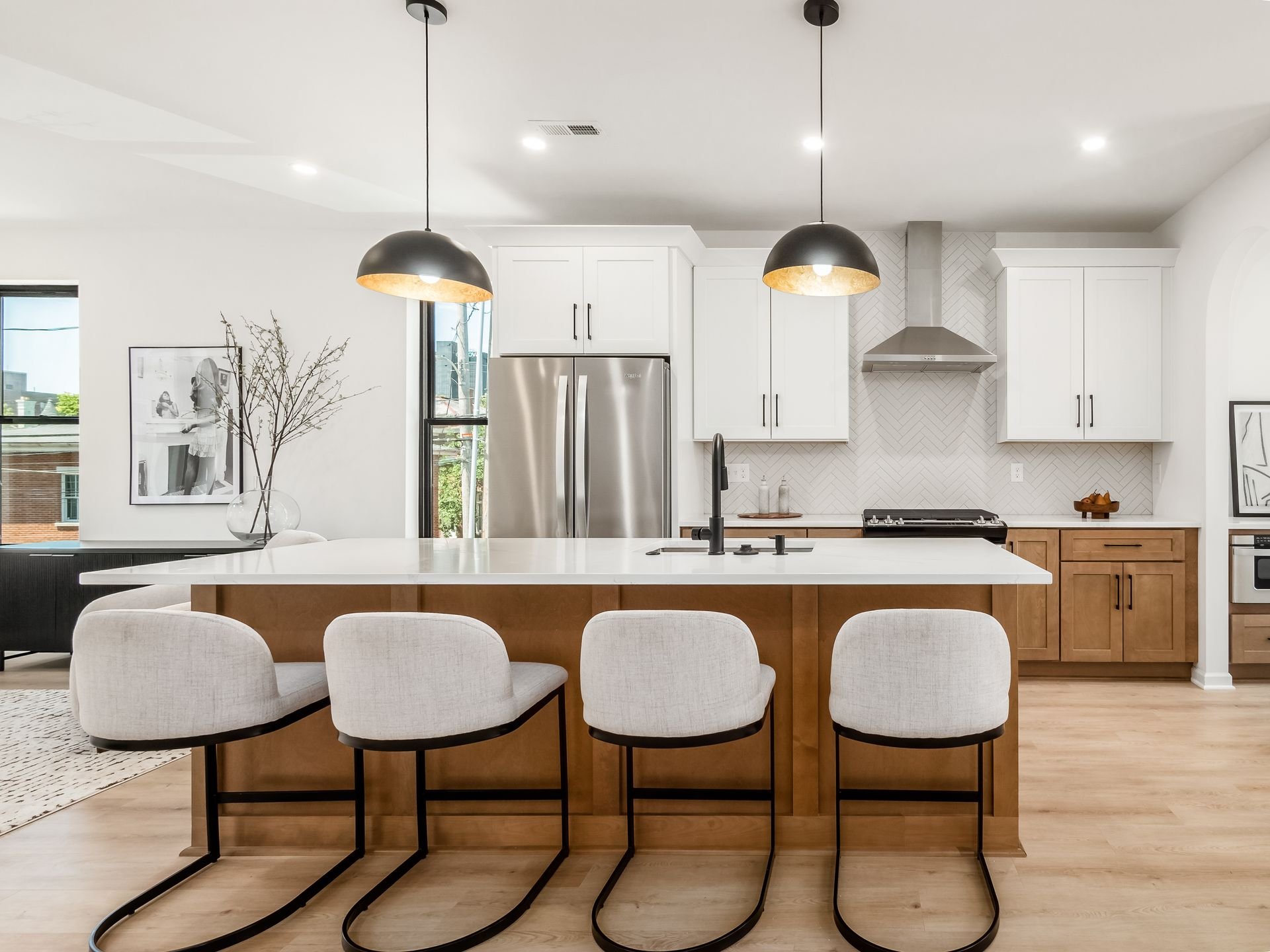 Modern kitchen interior with white cabinets, stainless steel appliances, and a large island with four upholstered stools. Overhead are two black pendant lights. The backsplash features a herringbone pattern.