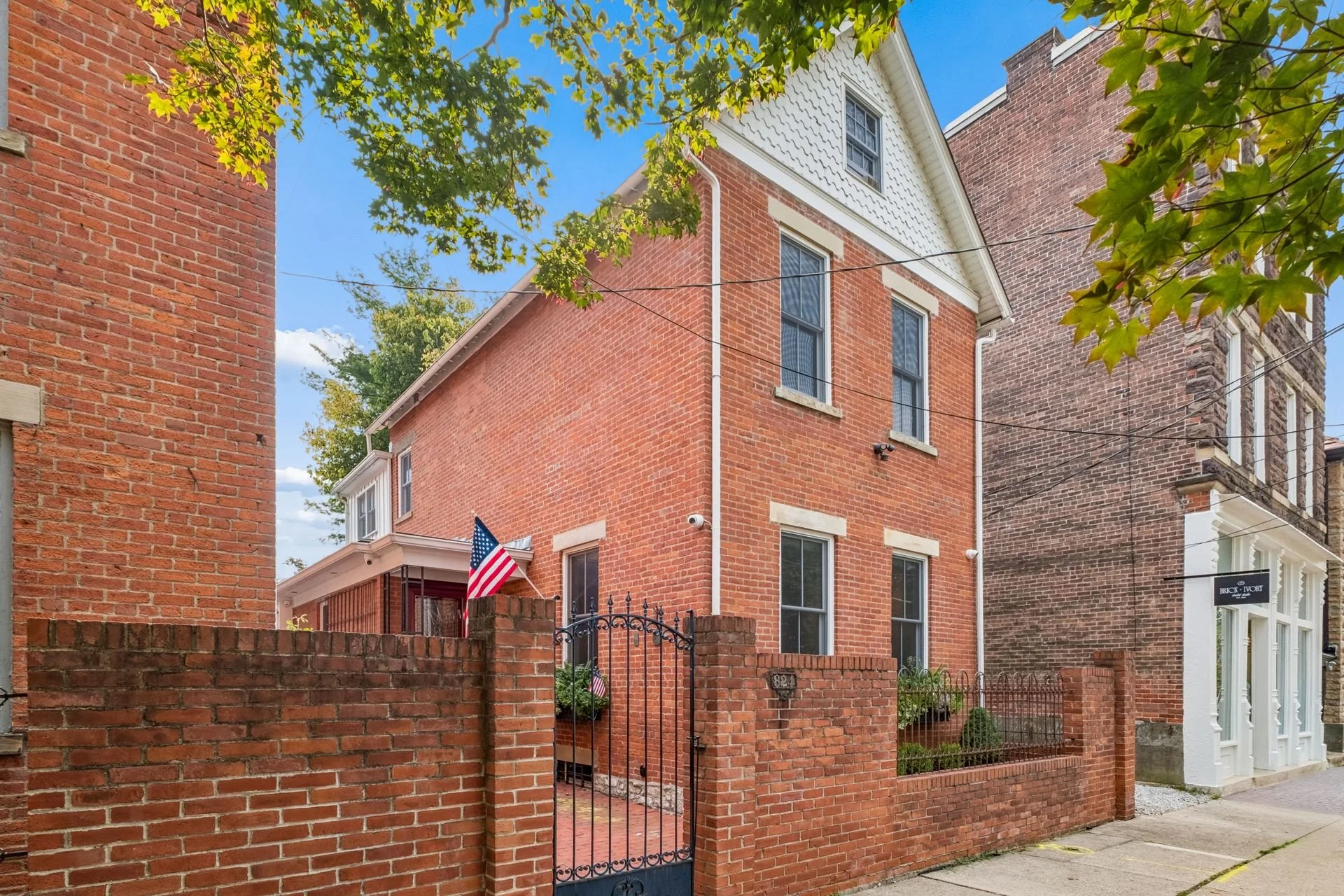 The image shows a brick house with a small front yard and a black iron gate. An American flag is hanging near the entrance. The house has three windows on the front and a partially visible second-story window. There are large green trees with yellow-