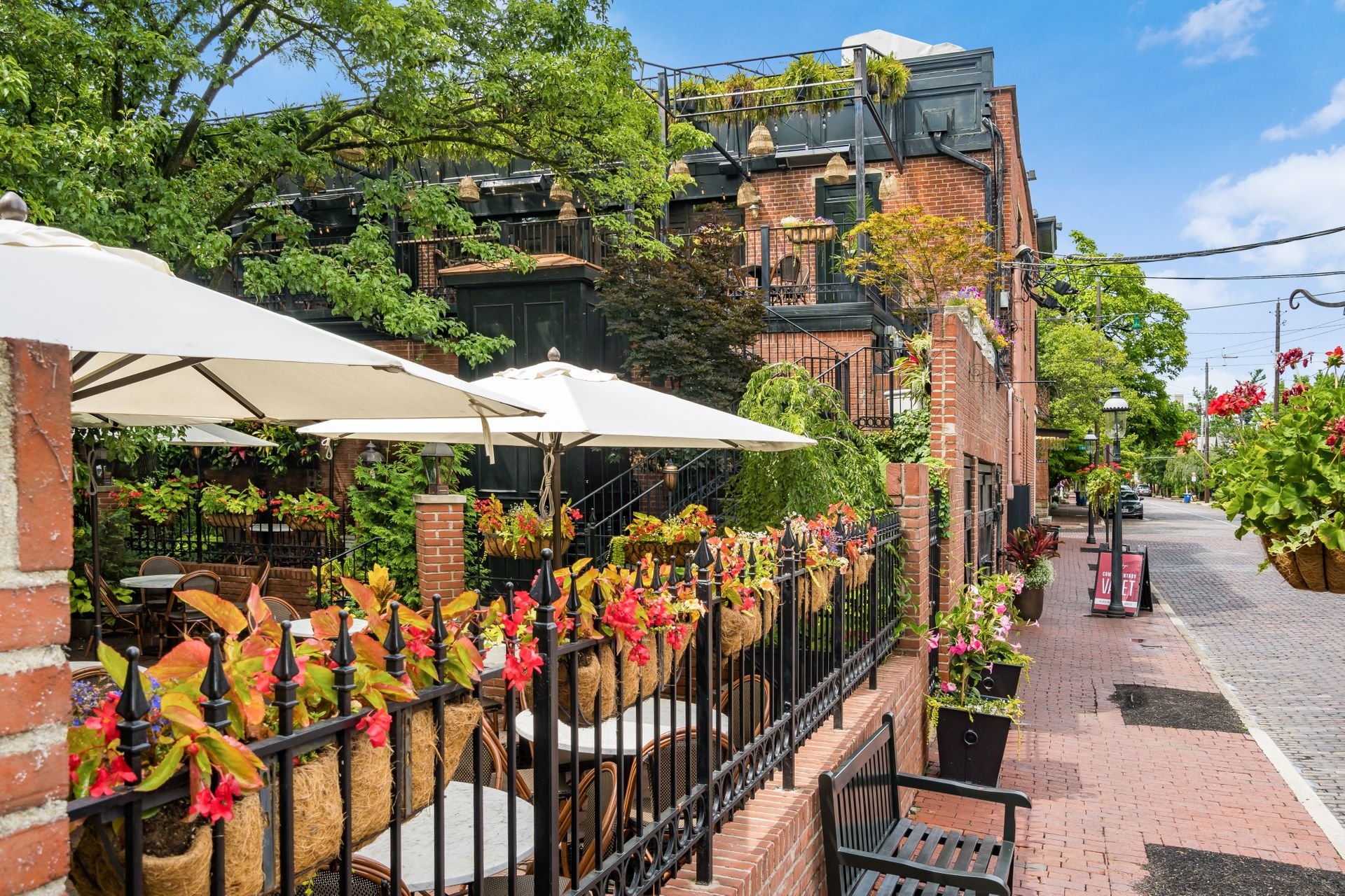 Charming outdoor cafe patio with tables and chairs under large white umbrellas, surrounded by colorful flowers and brick buildings, located on a cobblestone street with lush greenery.