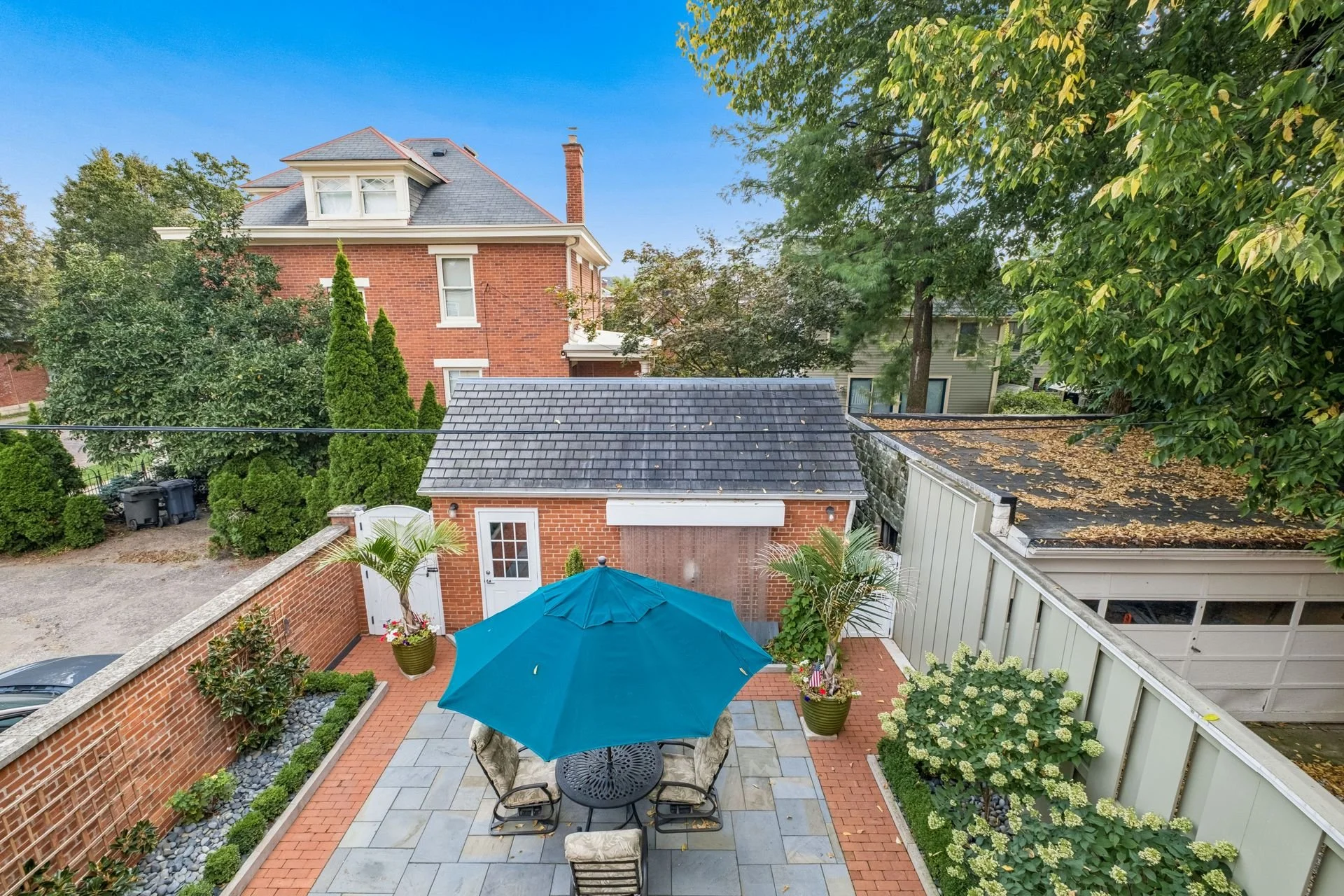 A backyard patio with a round table, six chairs, a blue umbrella, potted plants, and a brick wall. The patio is paved with grey and red bricks, and the backyard is surrounded by greenery and trees. There is a small building with a garage door on one 