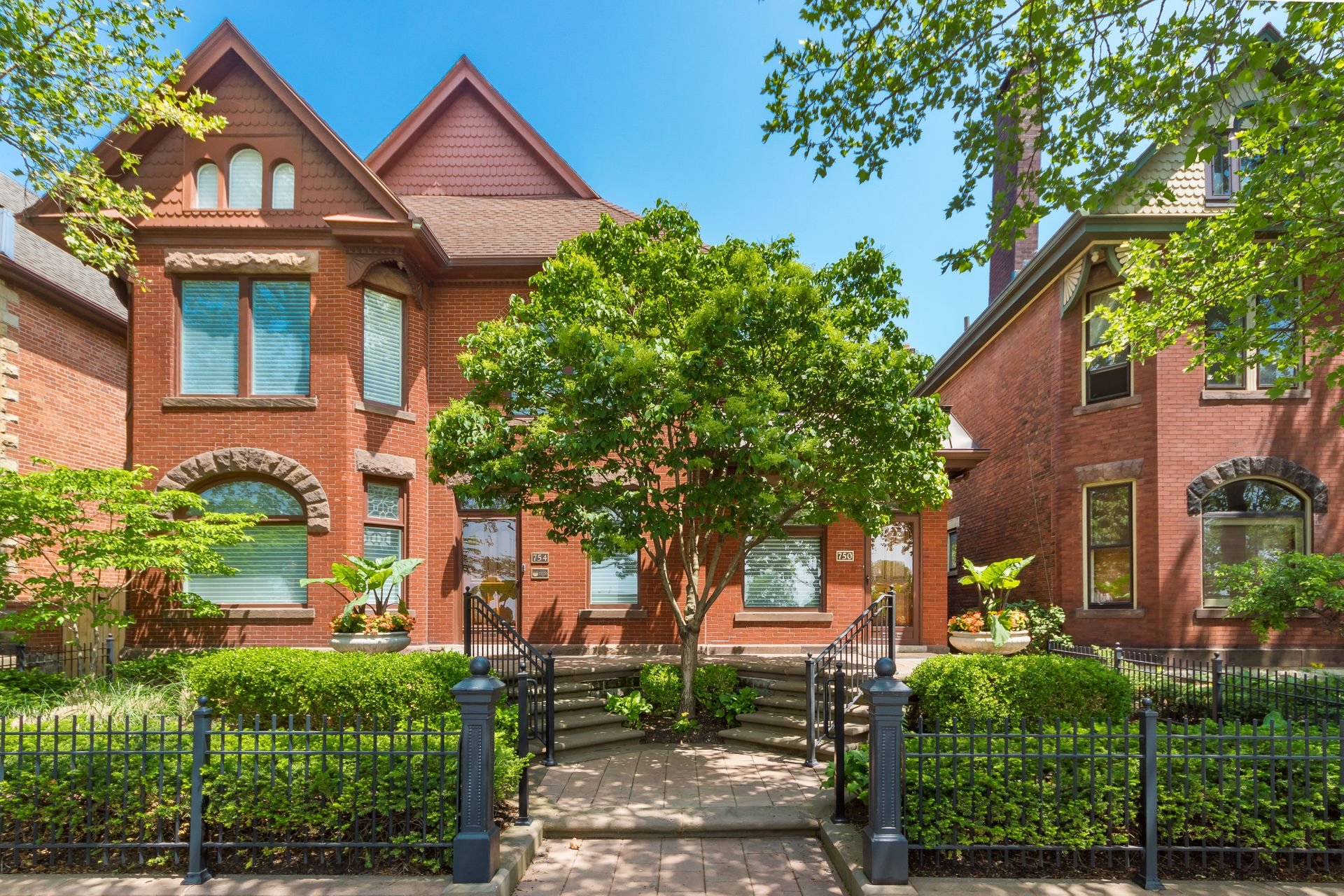 A historic red brick house with arched windows and decorative stone accents, surrounded by lush green trees and shrubs, set behind a black iron fence.