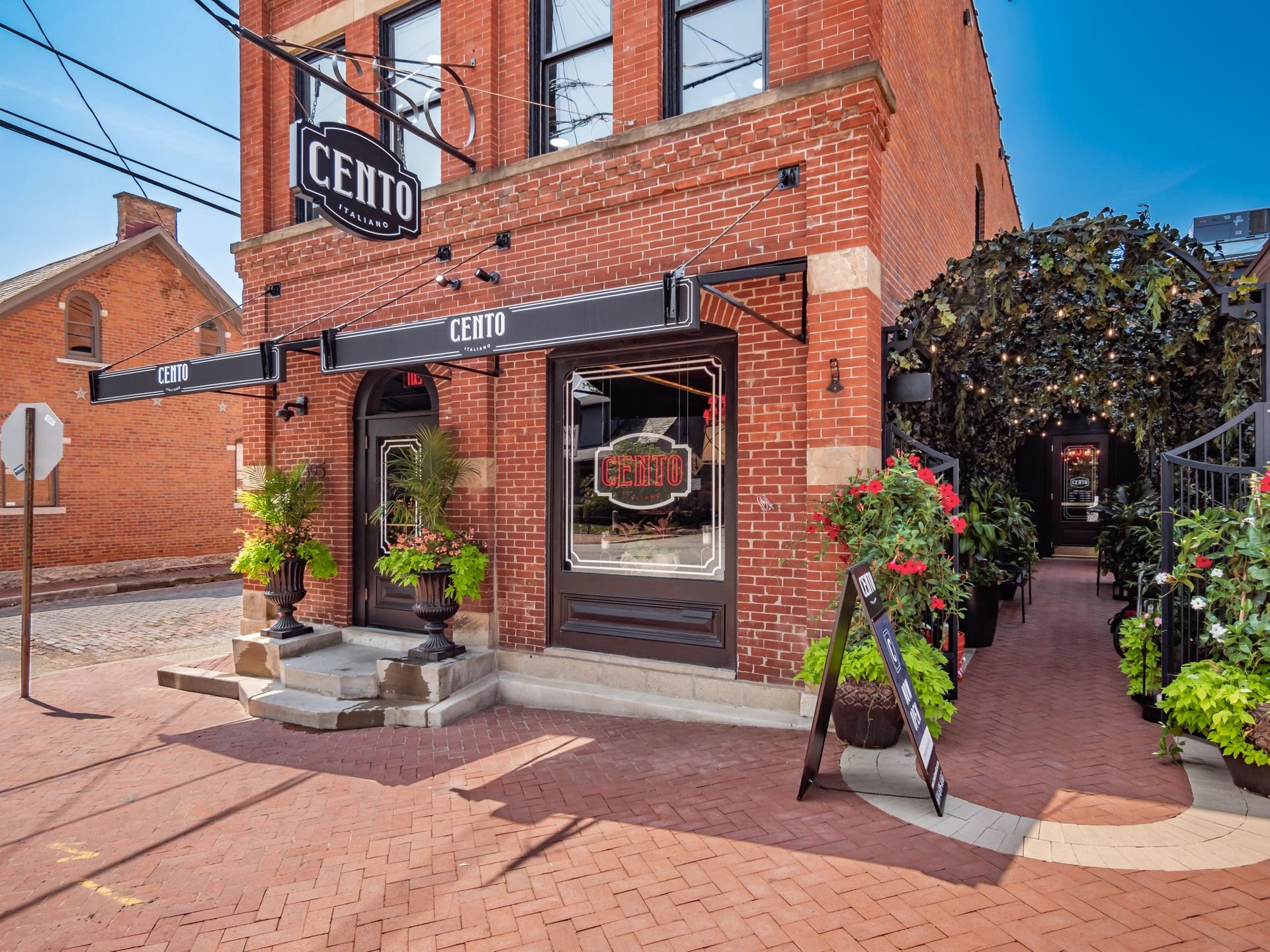 A brick building with a restaurant entrance labeled "CENTO" in large letters. The facade features a black awning and decorative plants in large pots on the doorstep. There's a covered outdoor walkway with greenery and string lights leading to an inte