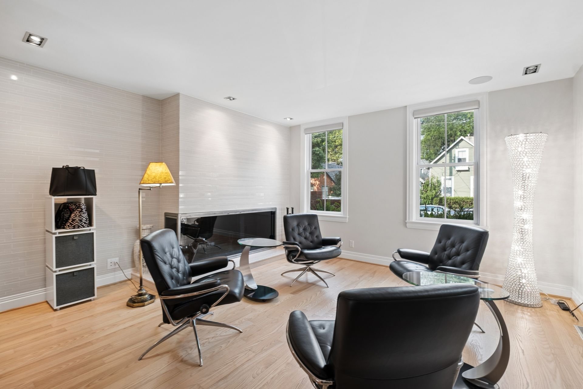 Modern living room with four black leather chairs around glass tables, large windows with white blinds, a white textured wall, a white lamb lampshade, and a white shelving unit with a black bag and patterned items.