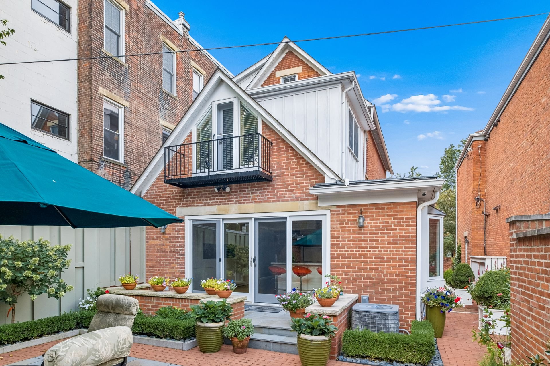 Backyard patio with potted plants, outdoor furniture, and a small balcony on the second floor of a brick house under a blue sky.