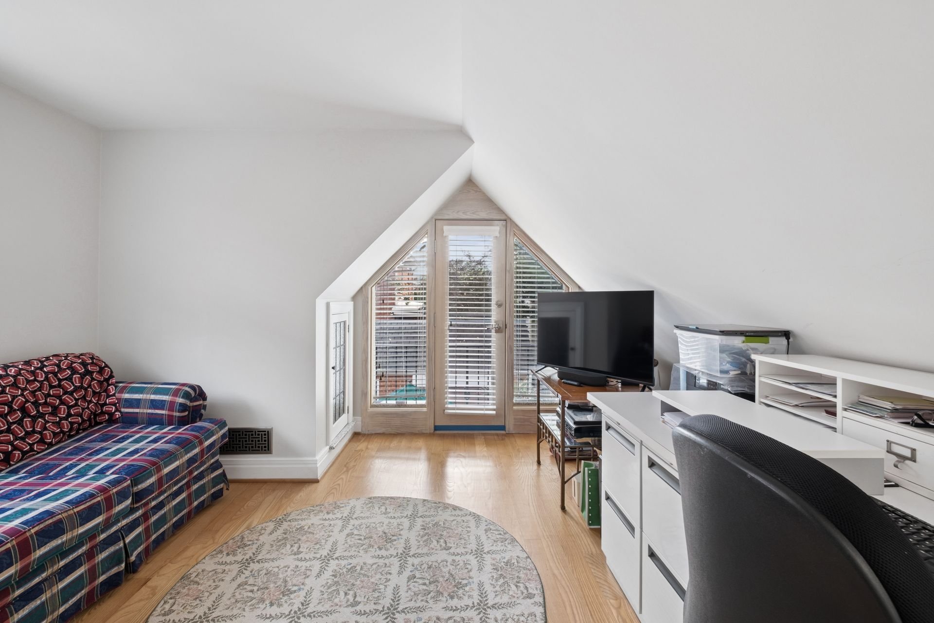 Living room with white walls and sloped ceiling, a plaid couch with football-themed fabric, a small white round rug, a black flat-screen TV on a wooden stand near glass doors with blinds, and a white shelf with books and office equipment.