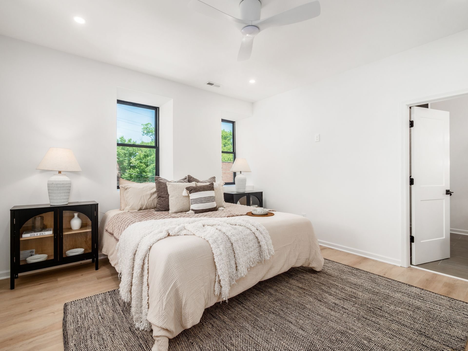 Modern bedroom with a large bed, neutral bedding, decorative pillows, and a cozy blanket. Two bedside lamps sit on dark nightstands with glass doors. Light wood flooring and a textured area rug. White walls and ceiling with recessed lighting, and a c