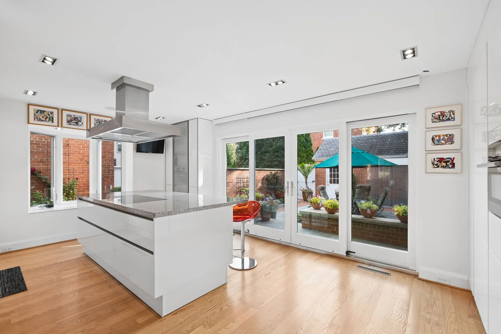 Modern kitchen with white cabinets, a grey countertop island, large glass sliding doors opening to a patio with outdoor furniture and green plants, and colorful abstract artworks on the wall.