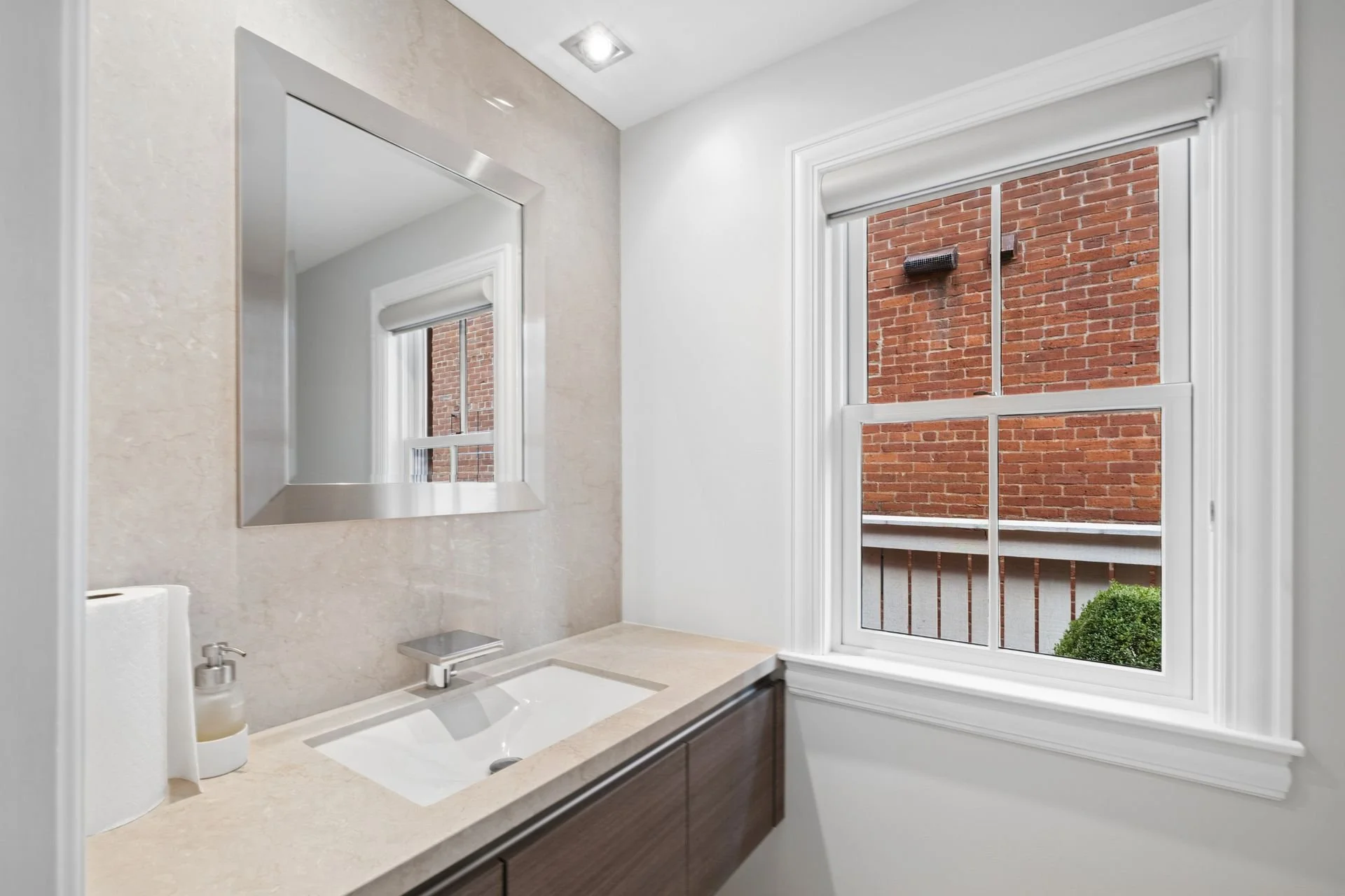 Bathroom with a mirror, sink, soap dispenser, and a window showing a brick wall outside.