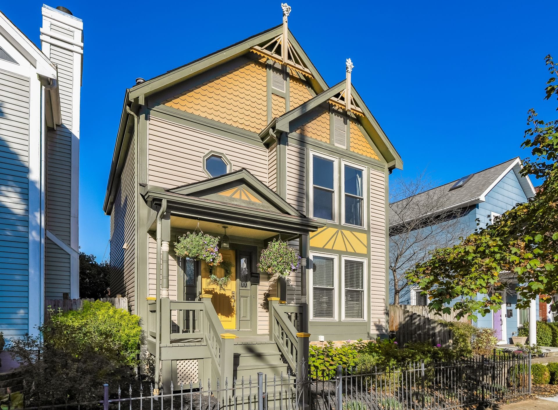 Victorian-style house with yellow and beige siding, gabled roof, and decorative trim, featuring a small porch with hanging plants and a picket fence in front.