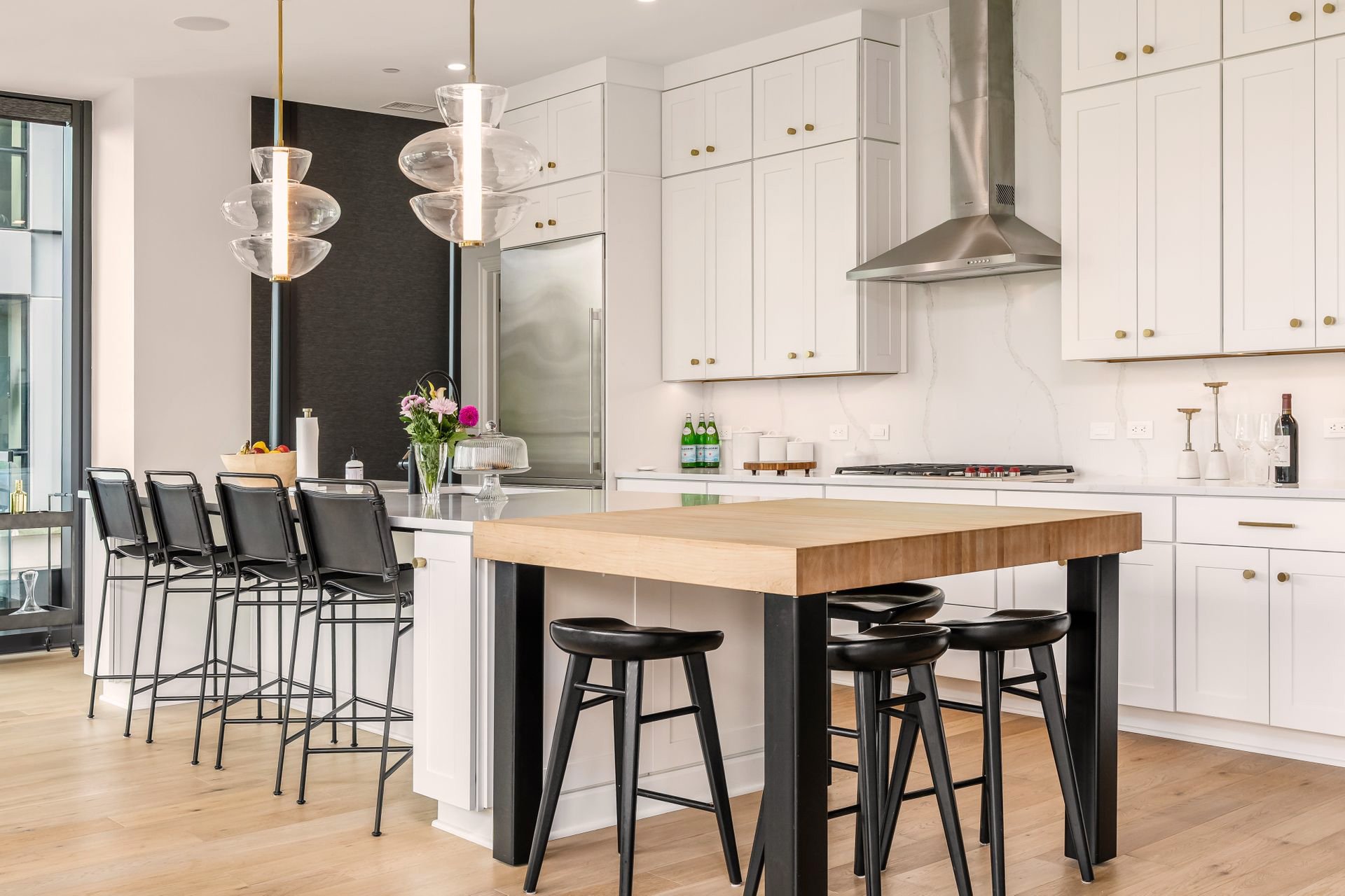 Modern kitchen with white cabinetry, wooden island, black stools, pendant lights, and stainless steel appliances.