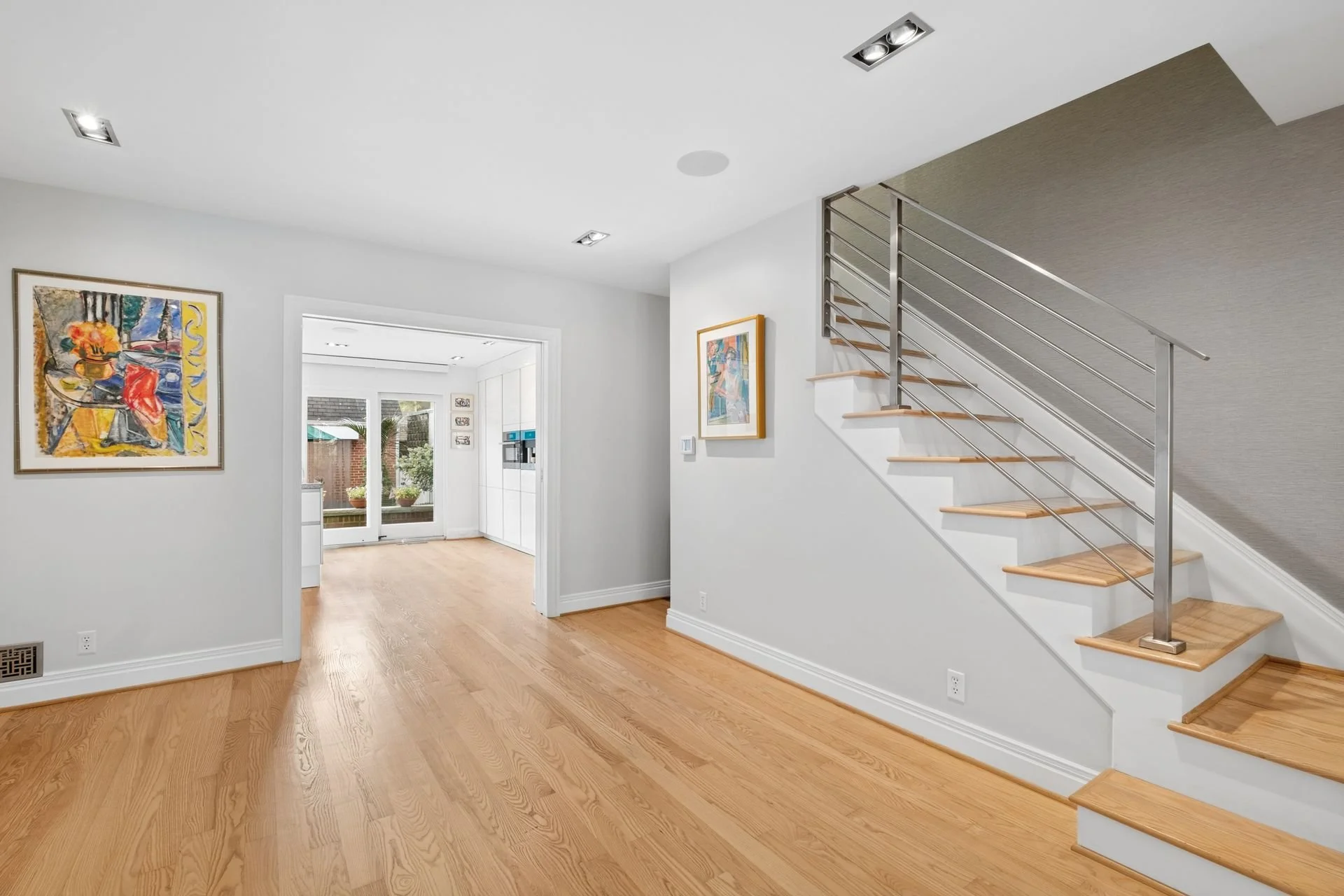 Interior view of a modern home with wooden floors, white walls, and artwork on the walls. A staircase with wooden steps and metal railings is on the right, leading to the upper level. In the background, there is a sliding glass door leading to an out