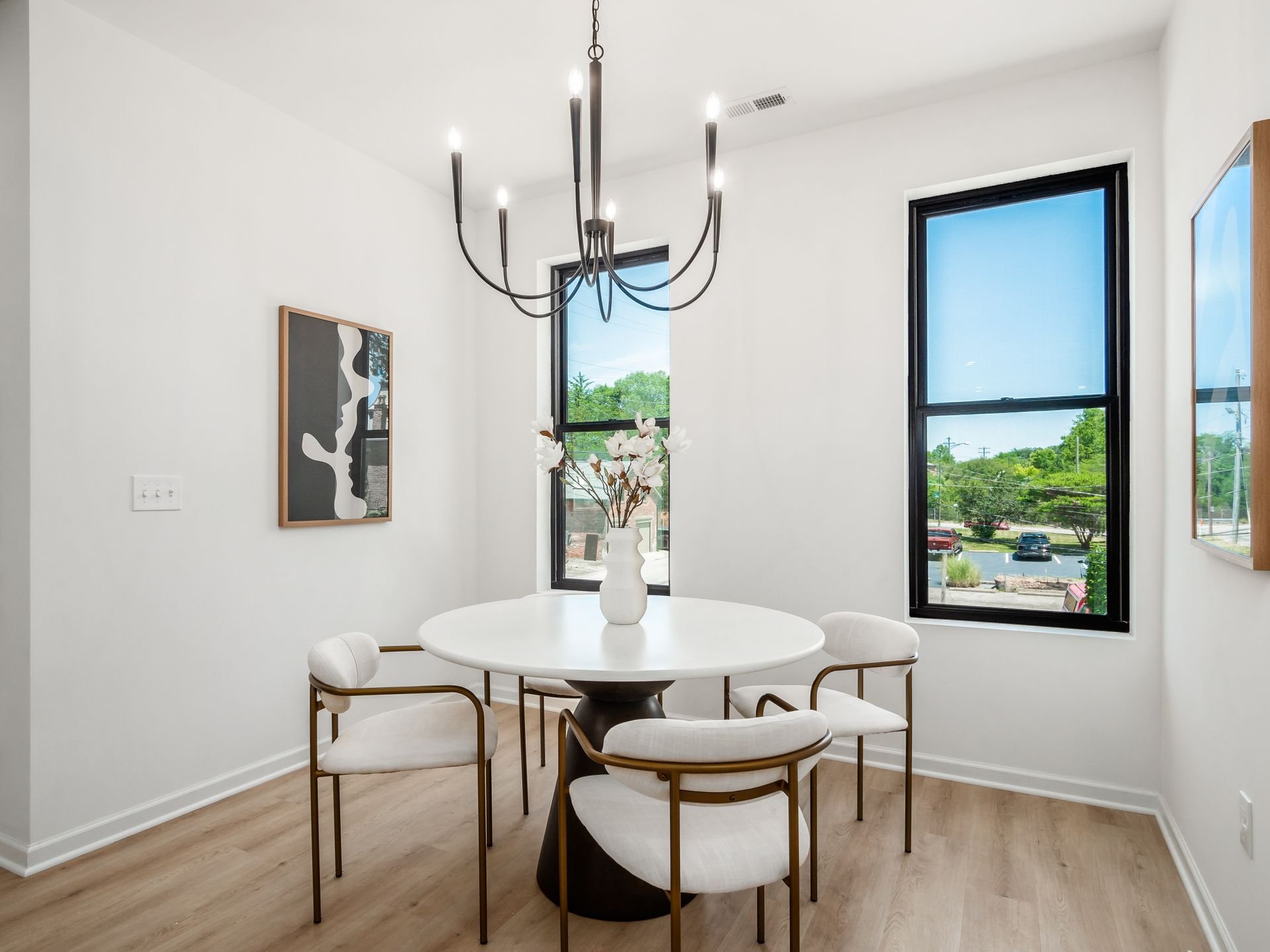 Modern dining room with round white table, four chairs with white upholstered seats, a black chandelier, and abstract wall art. Two large windows offer a view of greenery outside.