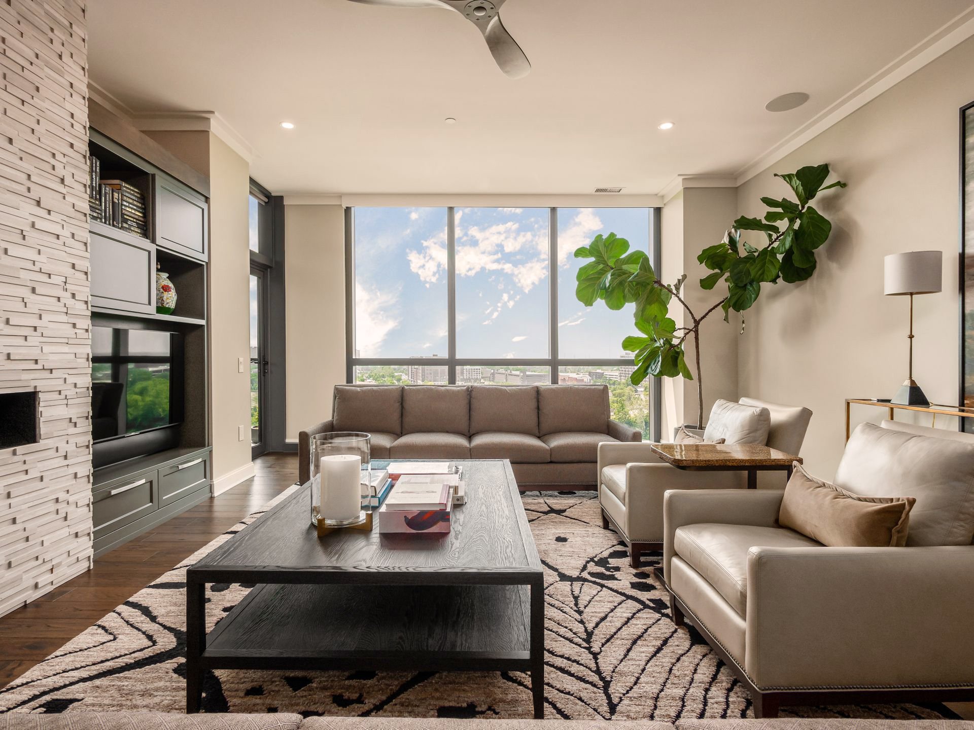Modern living room with large window, neutral-toned couch, armchairs, black coffee table, and a large potted plant indoors.