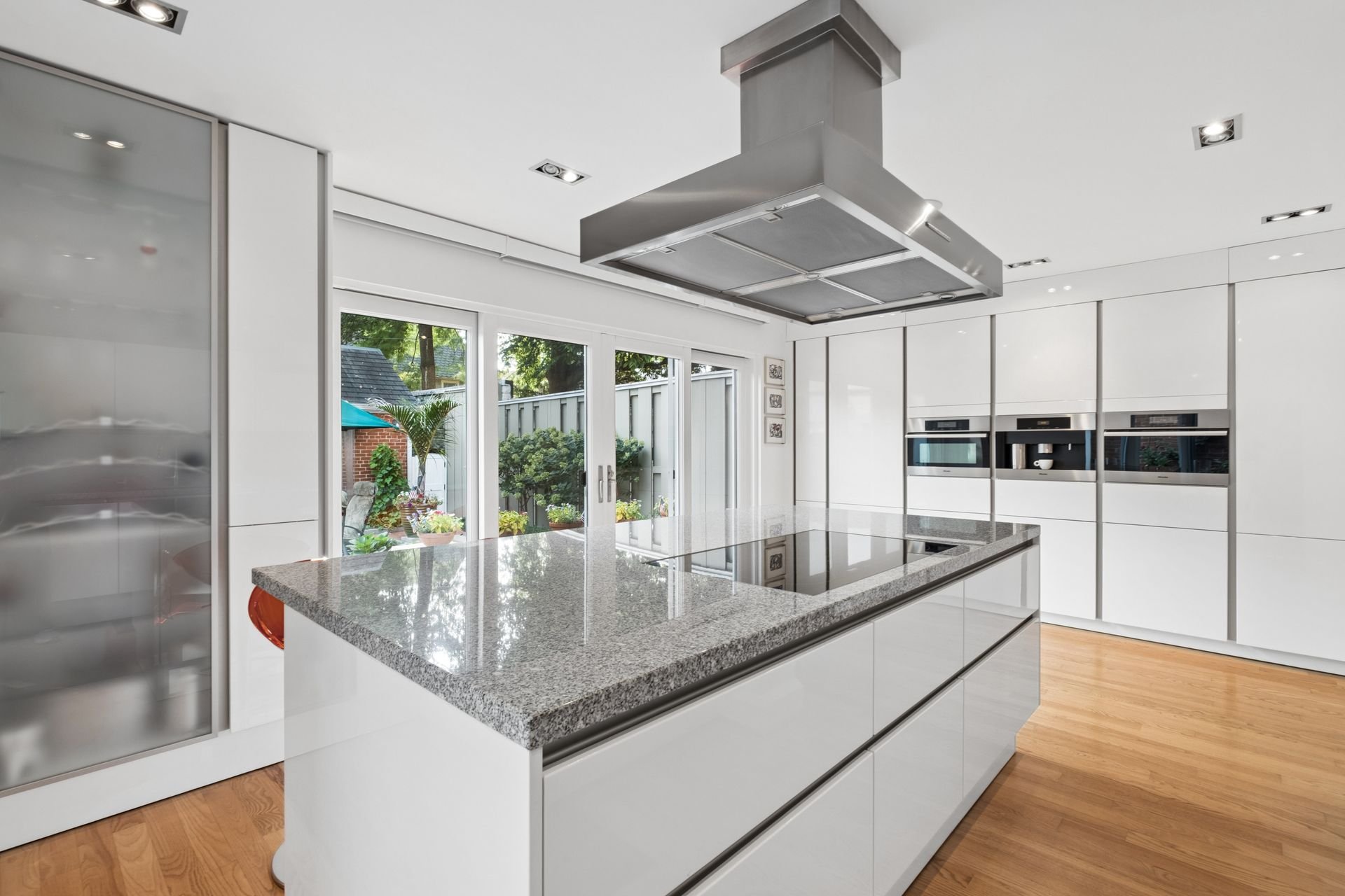 Modern white kitchen with large gray granite island, built-in ovens, and a stainless steel range hood, with large sliding glass doors opening to a backyard with greenery.