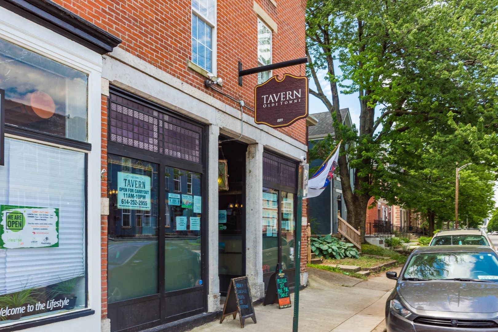 Street view of Tavern Olde Towne, a brick building with signs indicating it is open for carryout. A car is parked on the street, and trees line the sidewalk.
