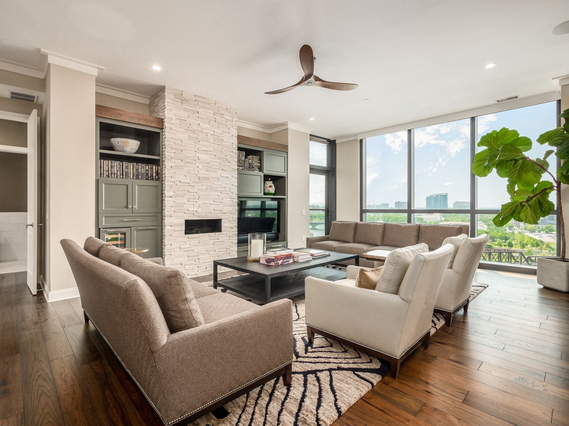 Modern living room with large windows, beige furniture, a stone fireplace, built-in shelves, and a ceiling fan. Hardwood floors and a patterned rug add to the decor.