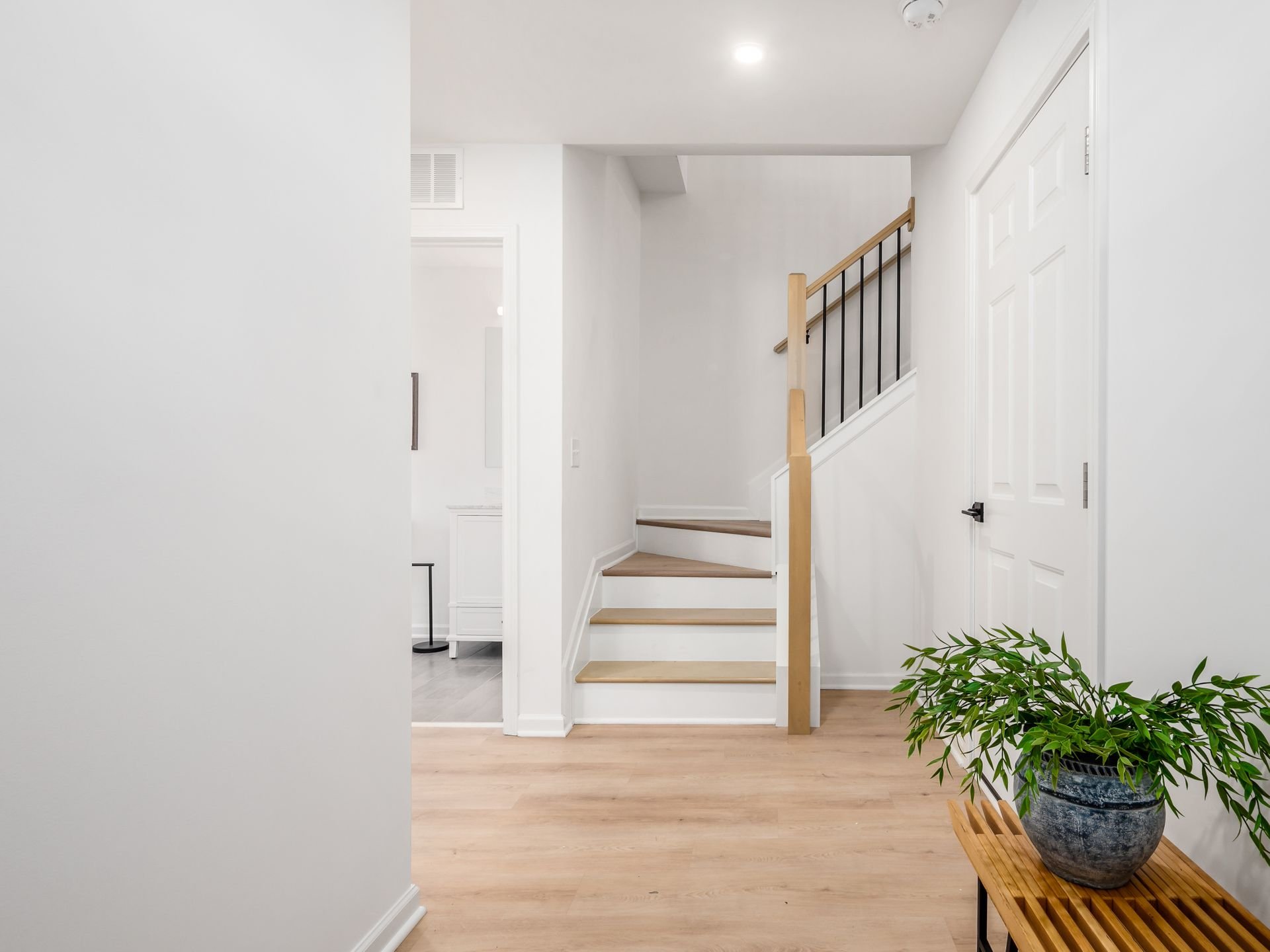 Modern, minimalist hallway with light wood flooring, featuring a small staircase with a wooden handrail. There's a small wooden bench with a potted plant on it near the stairs. The walls are painted white, and a partially open door is visible.