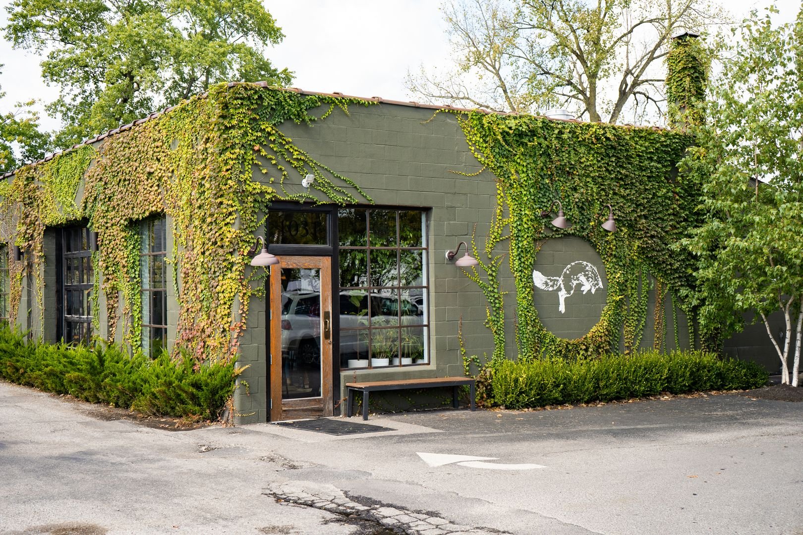 A green building with ivy-covered walls, featuring large windows and a wooden door. The exterior has a bench and decorative outdoor lights. A circular mural is partially visible, surrounded by greenery. Trees are in the background.