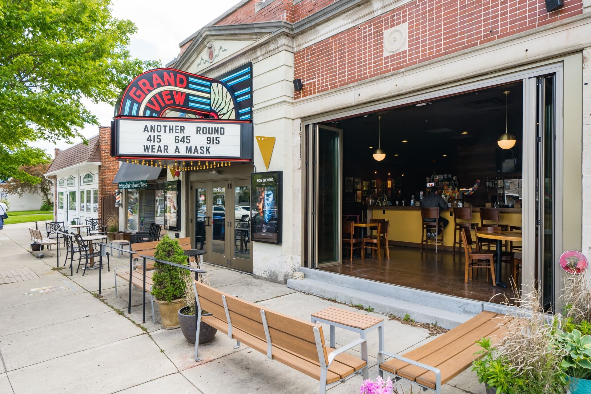 Exterior of a theater with an open cafe next to it, featuring a marquee displaying movie showtimes and a 'wear a mask' message.
