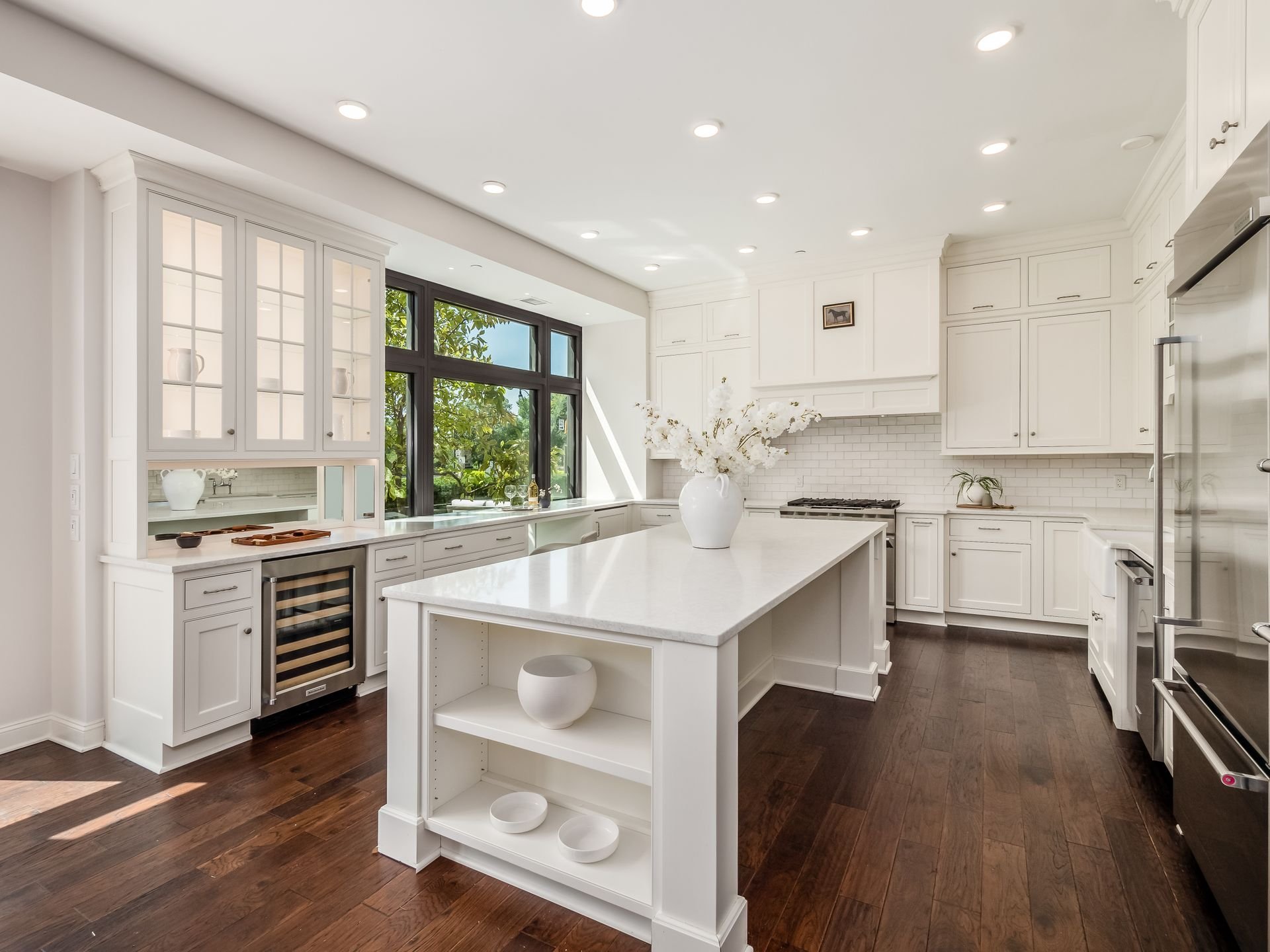 Modern kitchen with white cabinetry, large island with vase of flowers, hardwood floors, stainless steel appliances, and large windows.