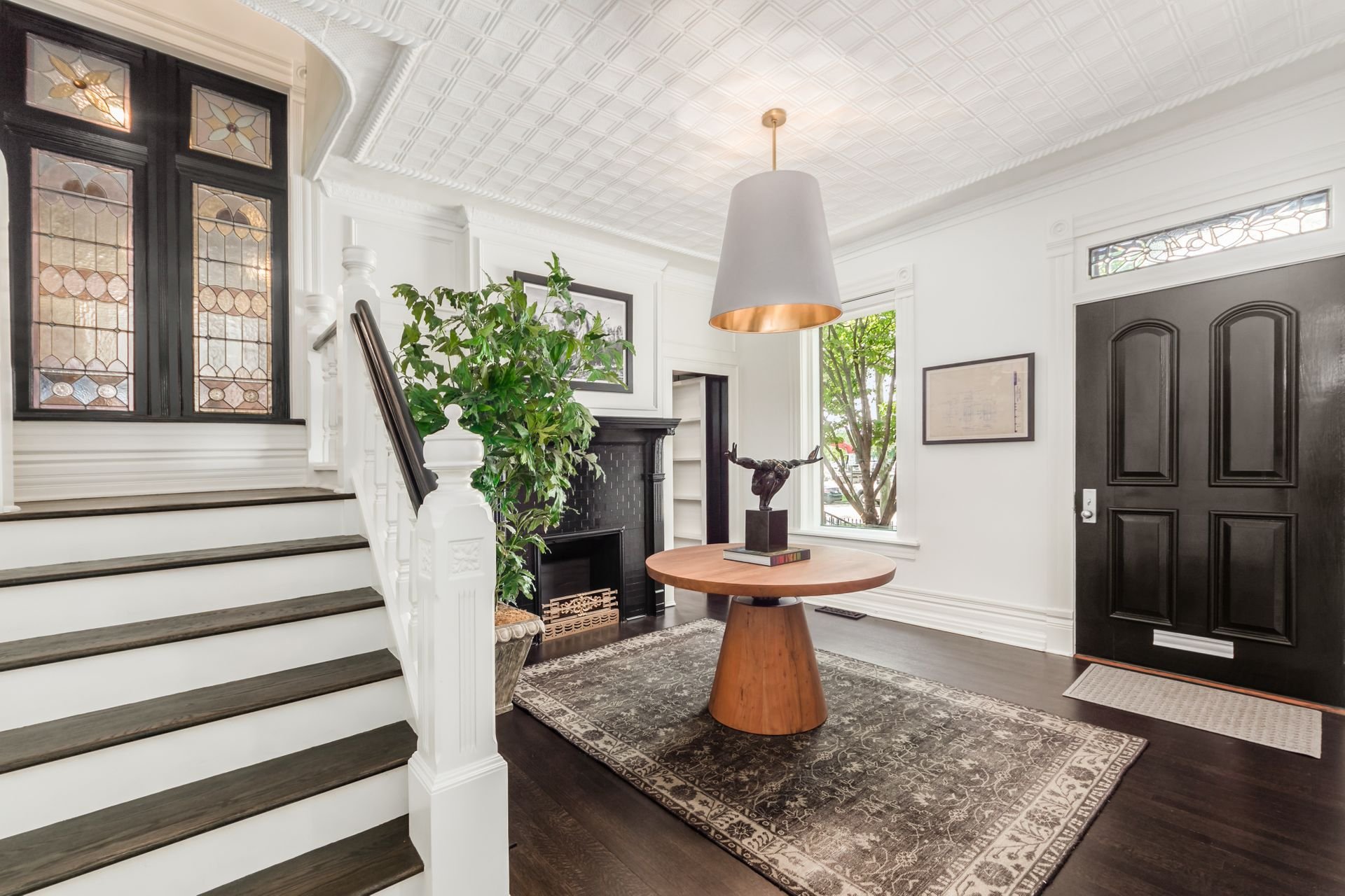 Elegant entryway with a staircase, stained glass windows, a round wooden table with sculpture, large potted plant, decorative ceiling, and a black front door.