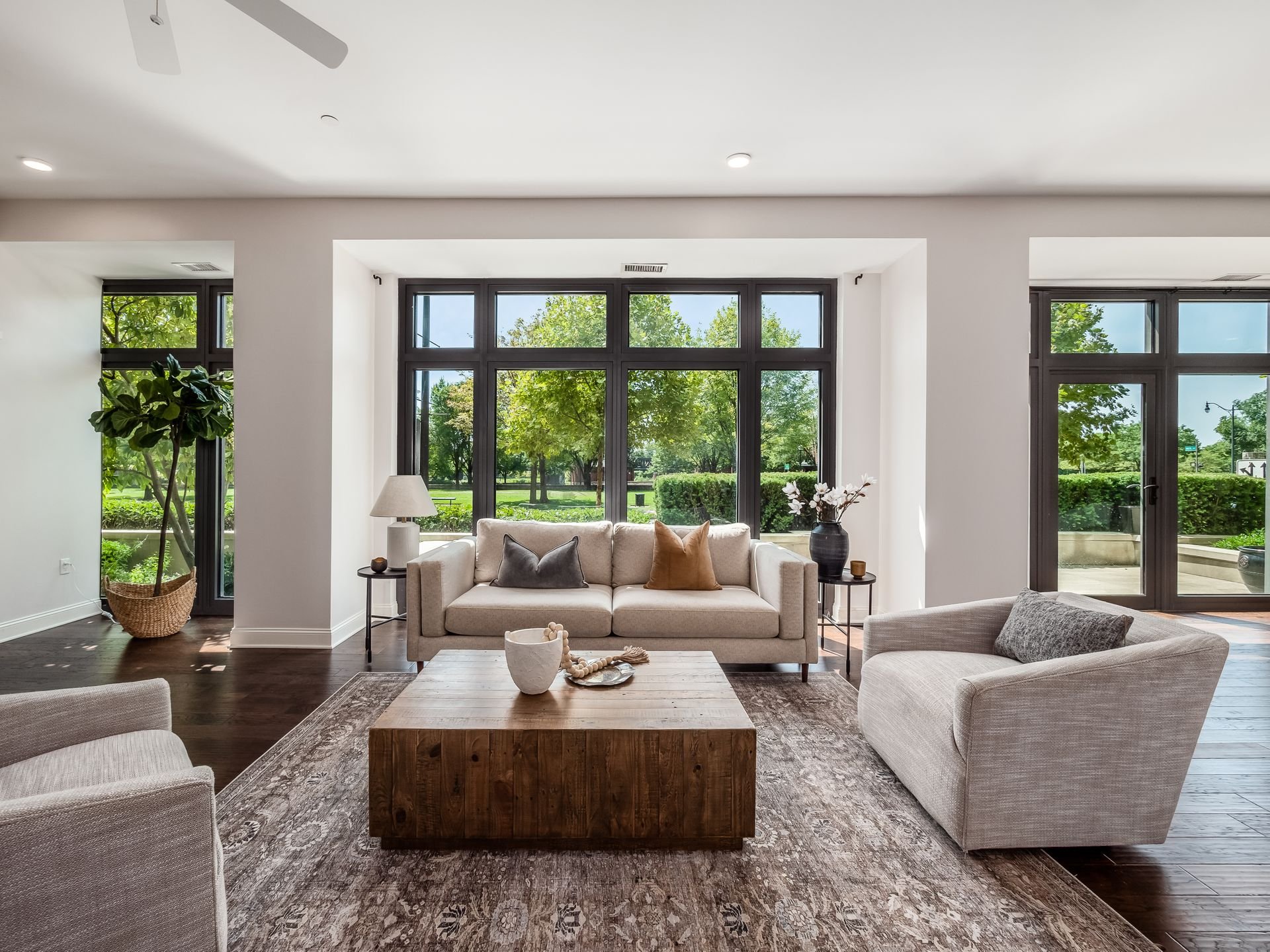 Modern living room with large windows, two beige sofas, a wooden coffee table, potted plant, and decorative cushions, overlooking a green garden.
