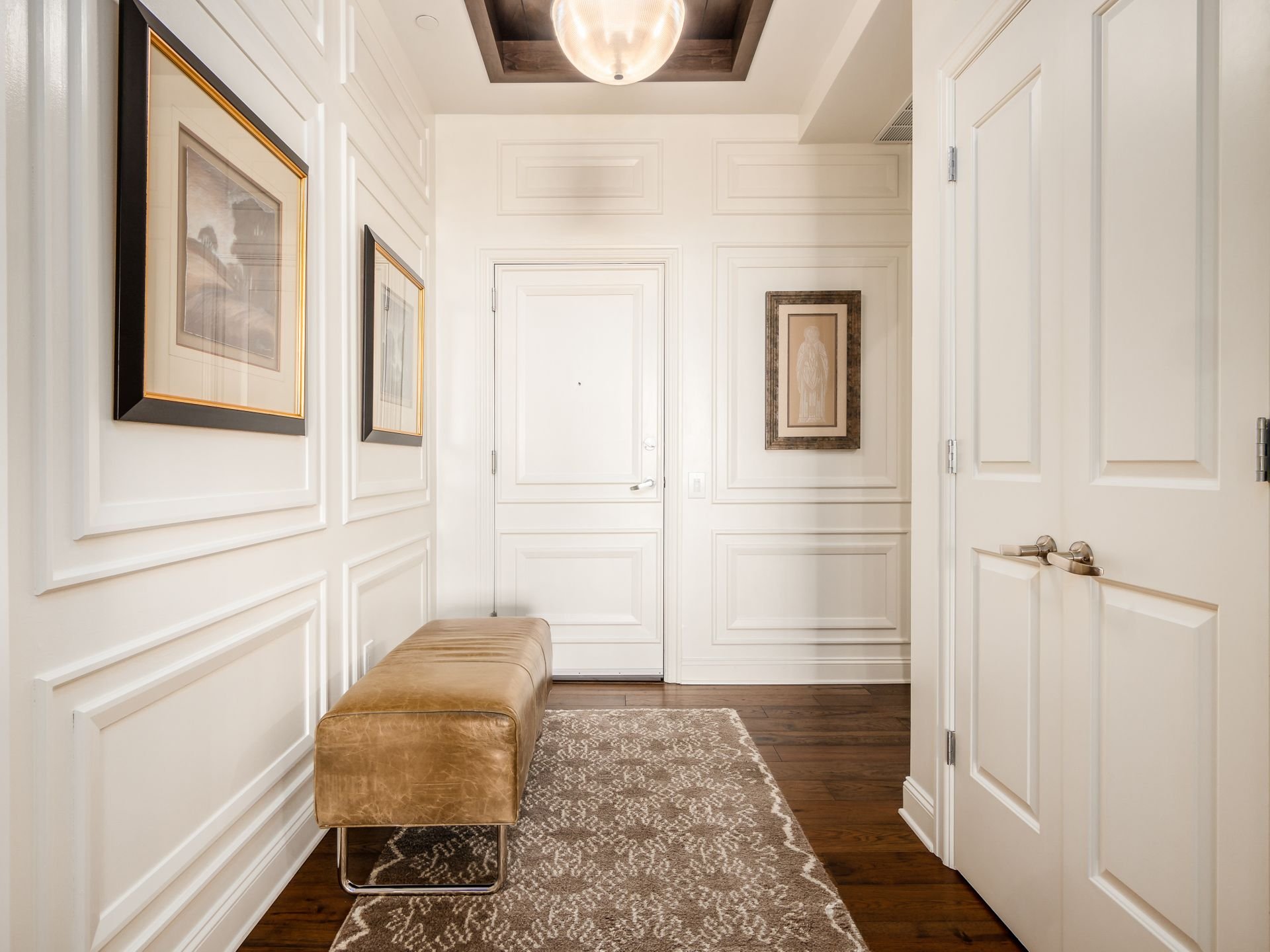 Elegant hallway with white paneling, framed art, a beige leather bench, and patterned rug on wooden floor.