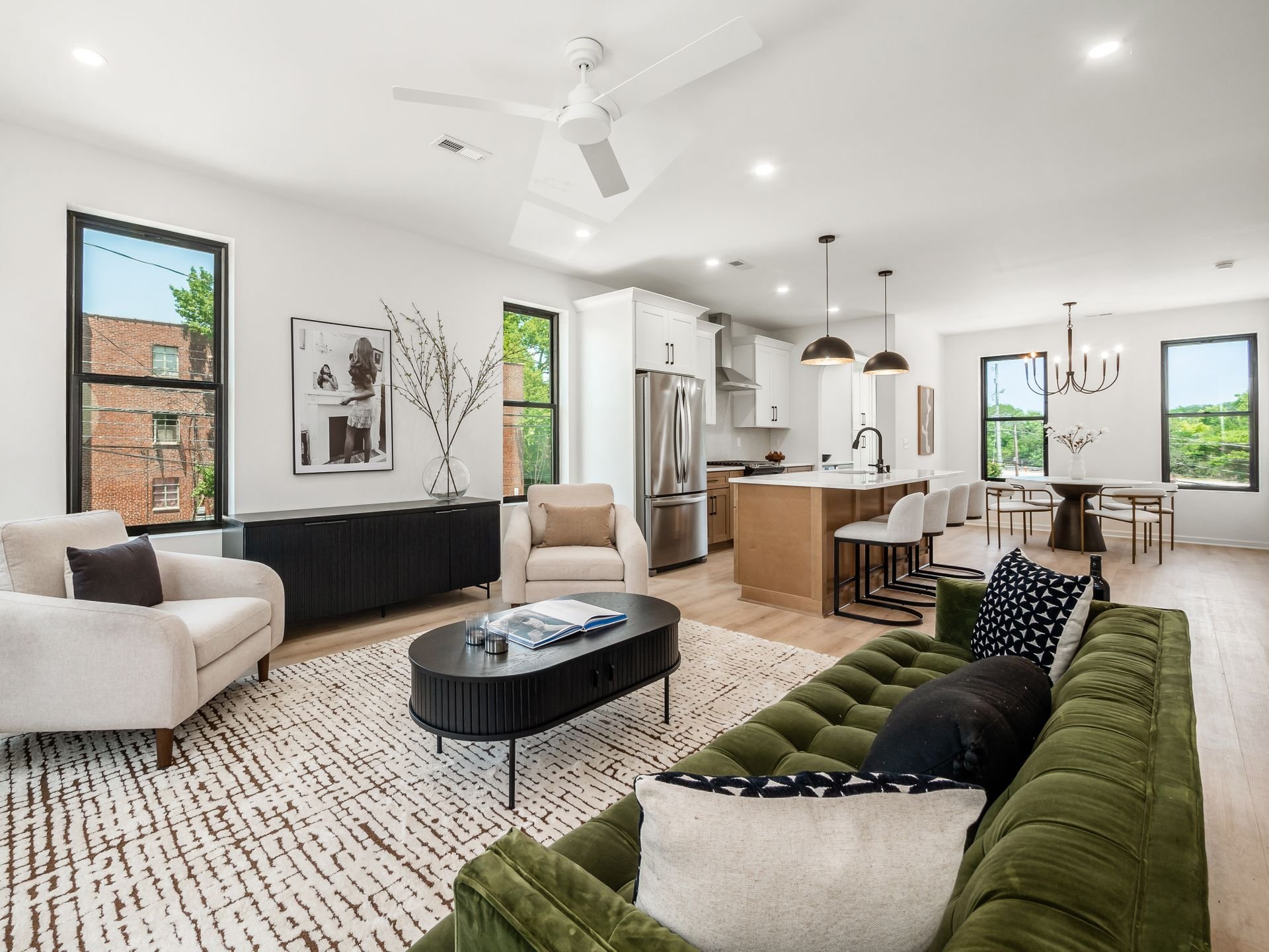 Modern open-concept living room and kitchen with white walls, black and white furniture, and wooden floors. Features an olive green sofa, a black coffee table, neutral armchairs, a textured rug, a kitchen island with stools, pendant lighting, and a d
