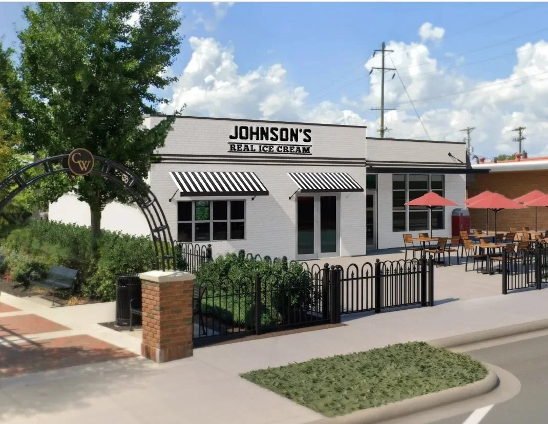 Exterior view of an ice cream shop named Johnson's Real Ice Cream, featuring a white brick building with black and white striped awnings, outdoor seating area with red umbrellas, and a landscaped entrance with a decorative arch.