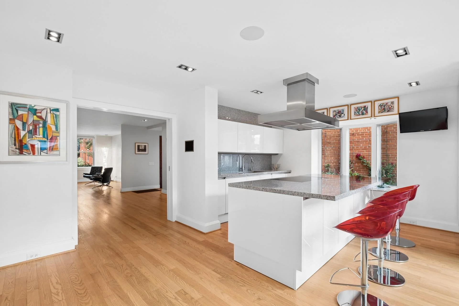Modern kitchen with white cabinets, gray countertops, and red bar stools. Bright space with hardwood floors and artwork on the wall.