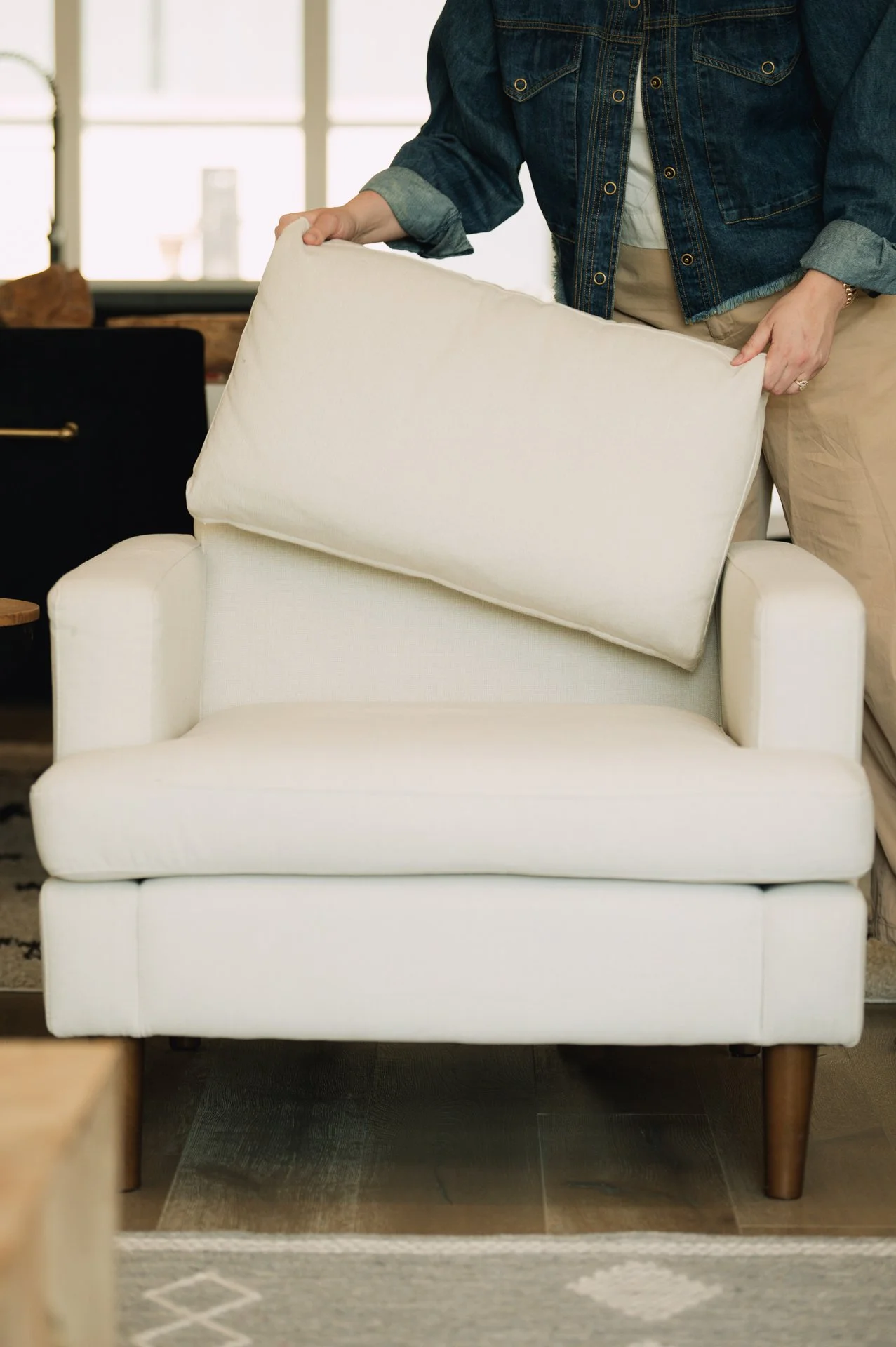 Person adjusting a cream-colored armchair with a cushion in a living room.