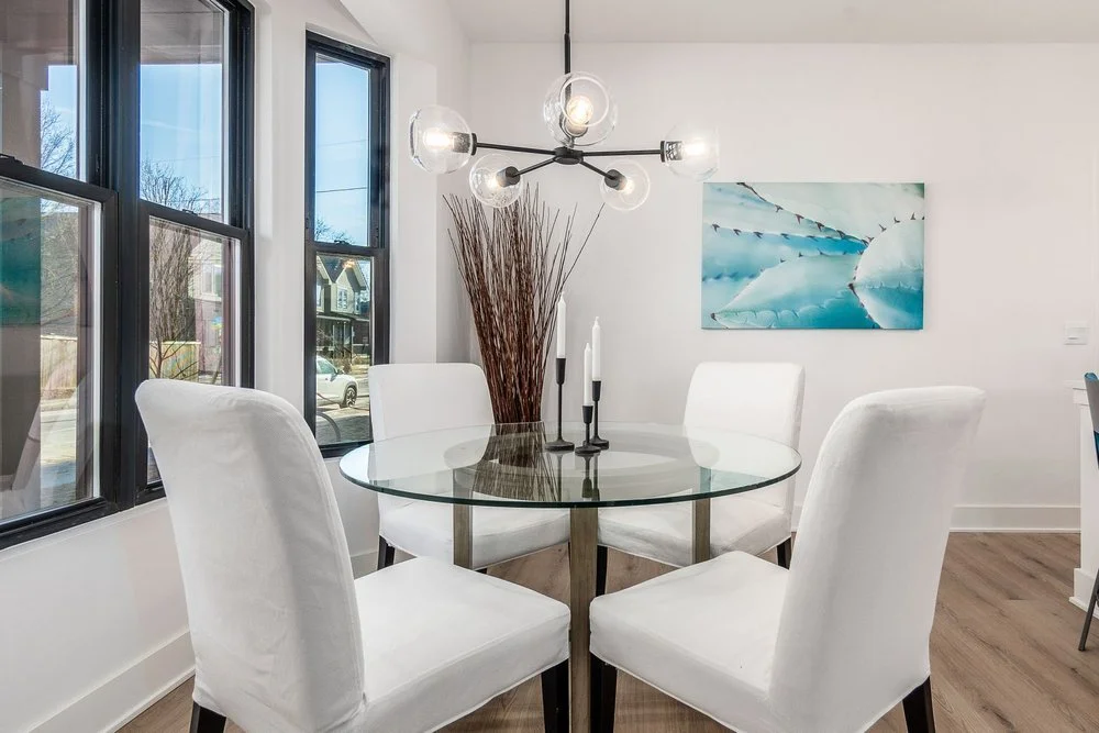 A modern dining room with a round glass table, four white upholstered chairs, a contemporary chandelier, a large window, and an abstract blue and white painting on the wall.