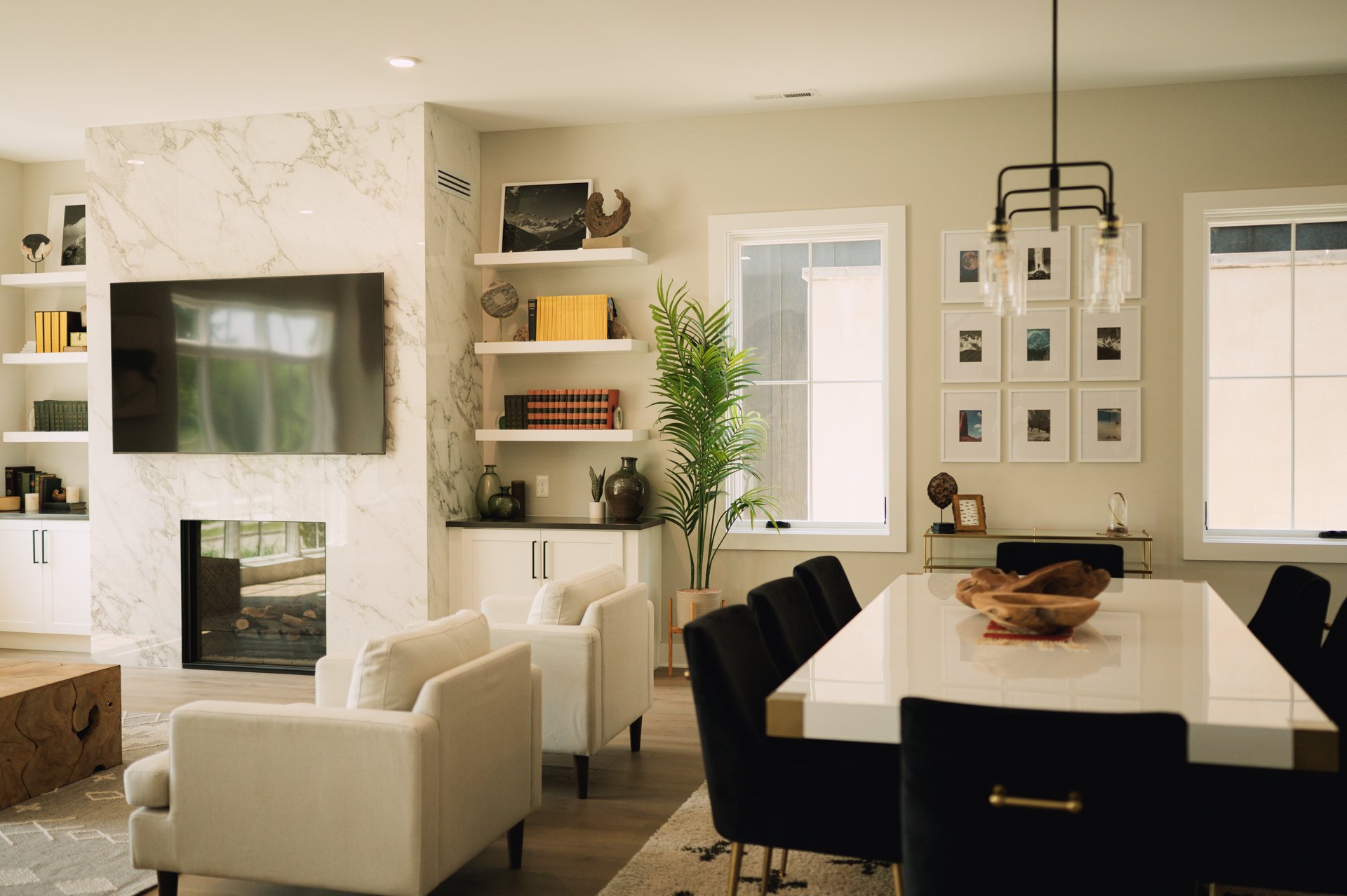 Modern living room with a marble fireplace and TV, white armchairs, black dining chairs, a large wooden coffee table, built-in shelves with books and decor, a tall potted plant, and framed photos on the wall.