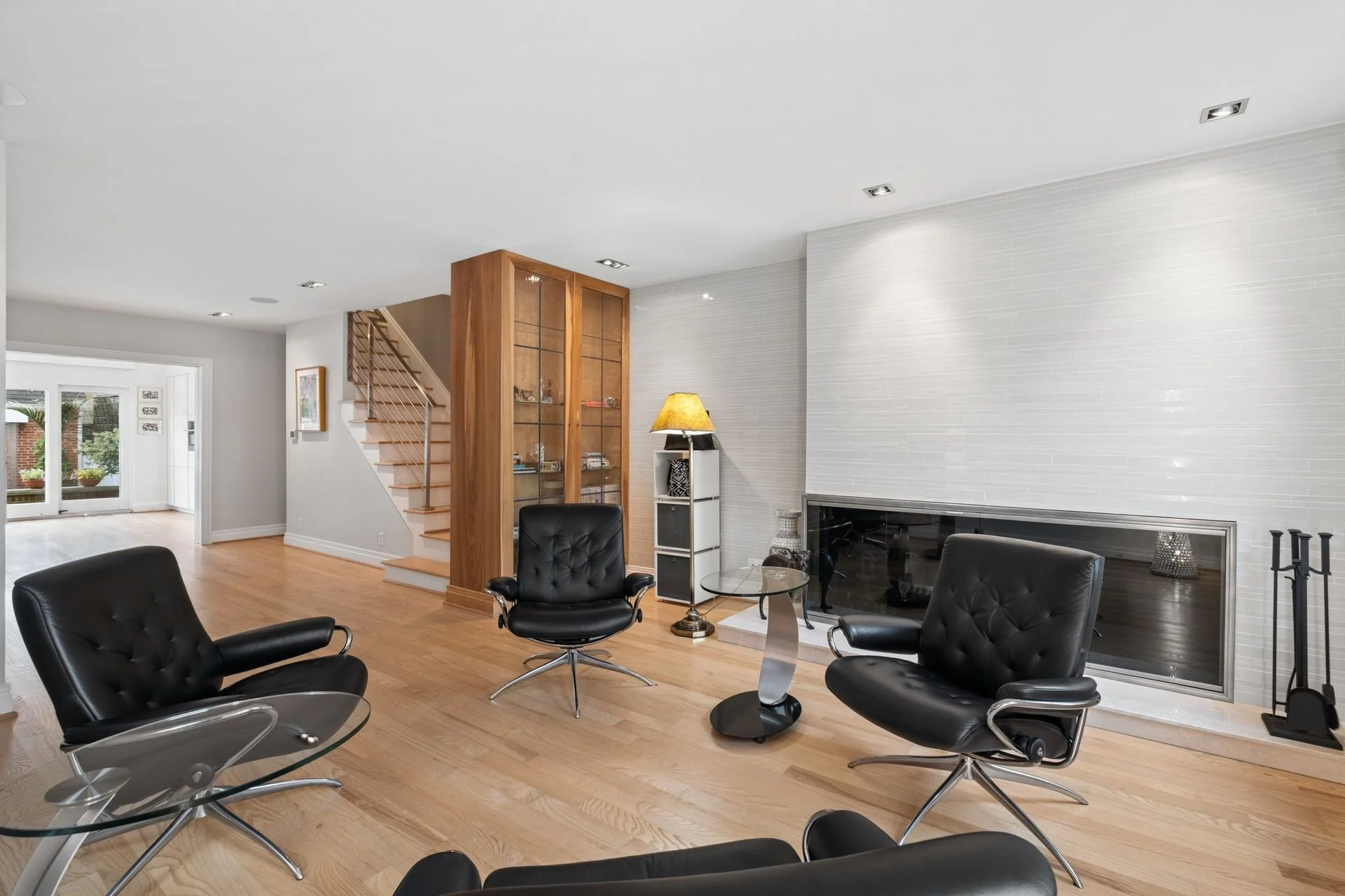 Modern living room featuring black leather chairs around a glass coffee table, a white brick fireplace, a wooden display cabinet, and a staircase with wooden steps and a metal railing, with natural light coming through sliding glass doors.