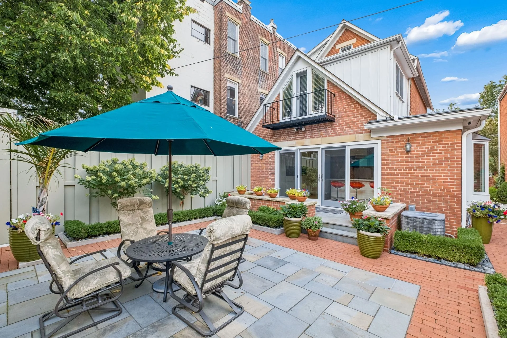 A backyard patio with a black metal dining table, six cushioned chairs, and a large blue umbrella. The patio is paved with large gray tiles and bordered by a brick pathway. Several large potted plants and flowers decorate the space. There is a red br
