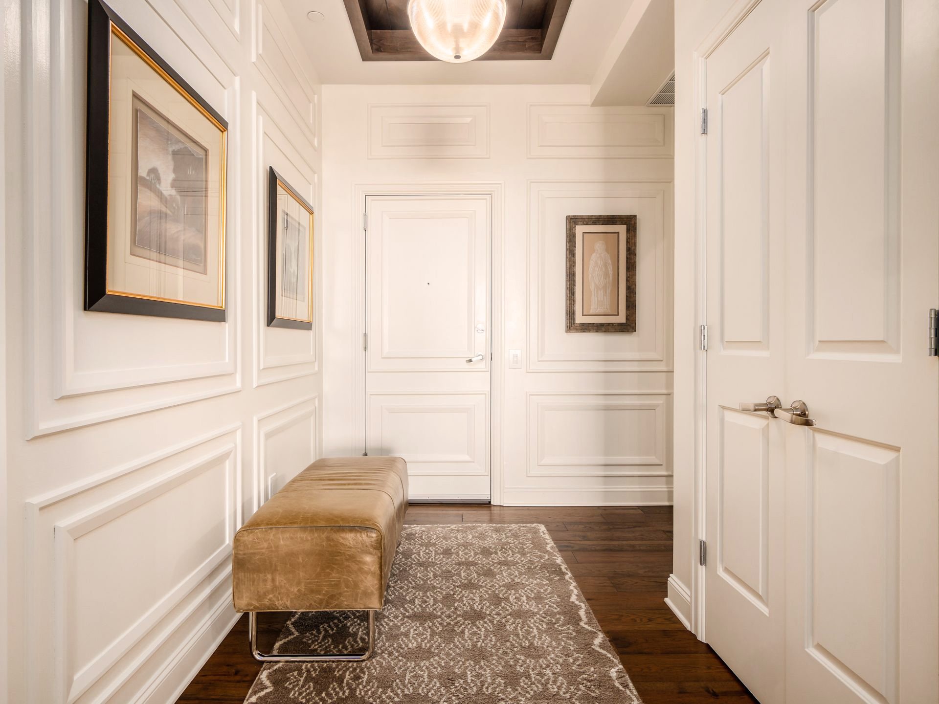 Hallway with cream-colored walls, wood flooring, upholstered bench, framed artwork, patterned rug, and a ceiling light fixture.