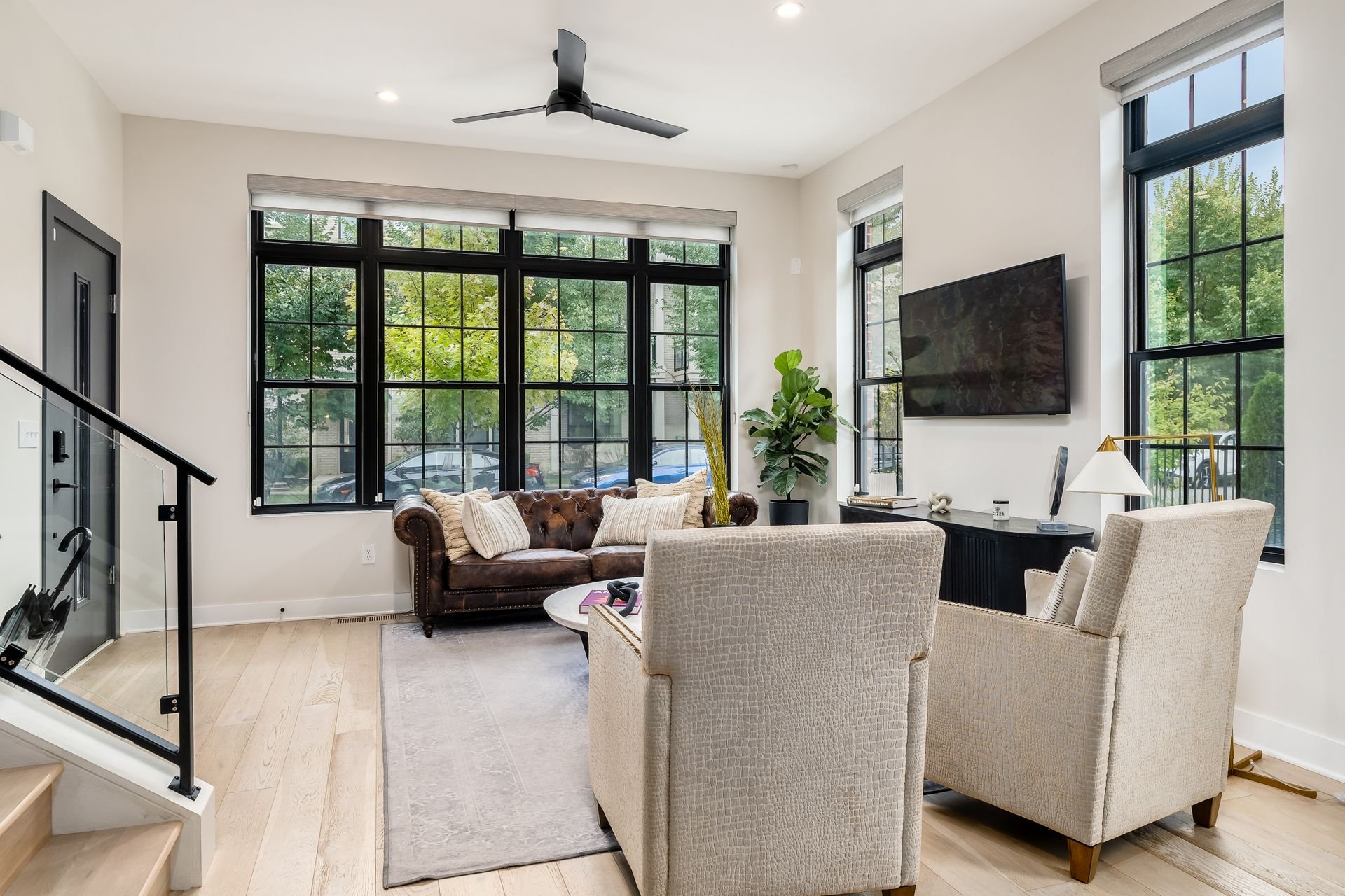 Modern living room with a brown leather sofa, two beige armchairs, large windows, a rug, flat-screen TV, and a potted plant.