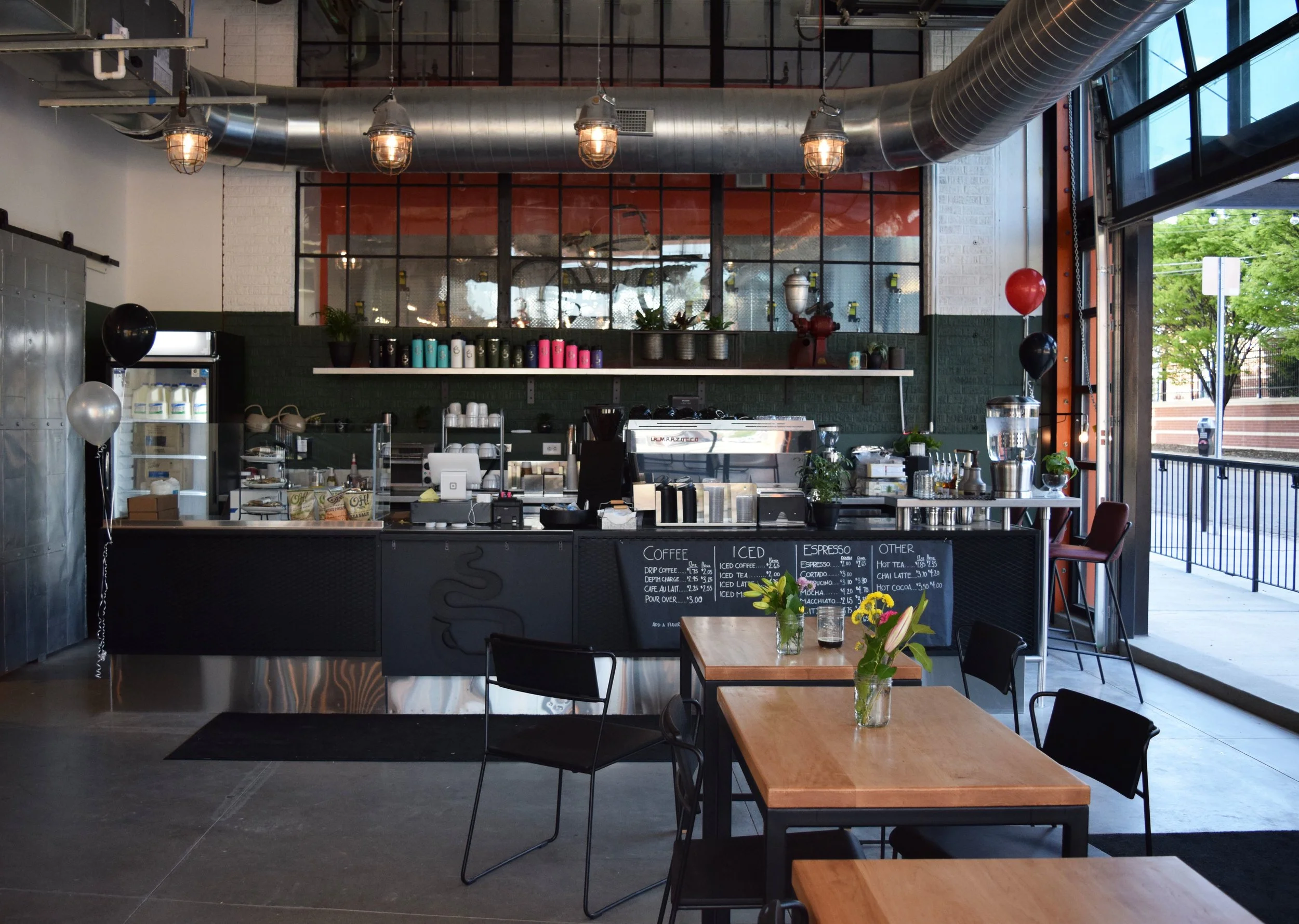 Interior of a modern coffee shop with industrial decor, featuring a counter with an espresso machine, colorful cups, chalkboard menu, and wooden tables with chairs. Balloons and potted plants adorn the area, and large windows provide an outdoor view.