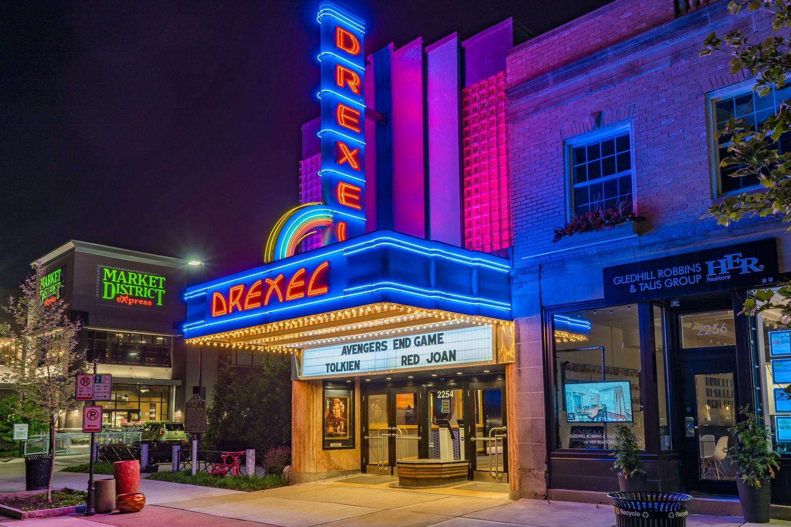 Drexel Theater with neon lights displaying movies; Market District Express sign nearby. Nighttime setting.