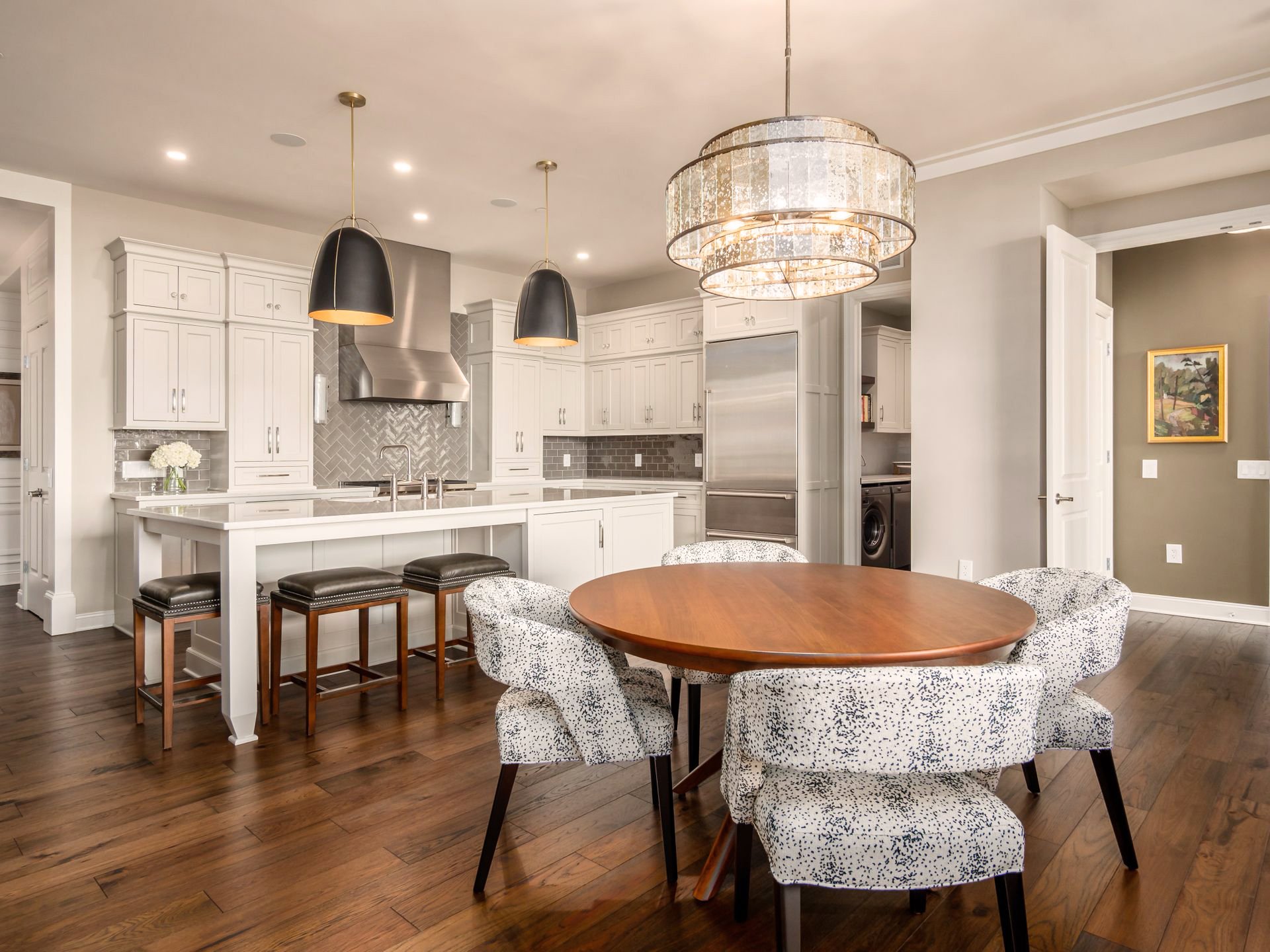 Modern kitchen with white cabinetry, stainless steel appliances, and an island with bar stools. A round wooden dining table is surrounded by patterned chairs. The room is illuminated by a large decorative chandelier and pendant lights.