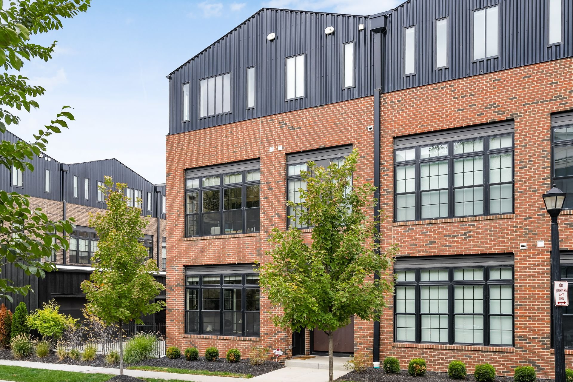 Modern brick townhouse with large windows, black metal accents, and landscaped front yard with small trees and bushes.