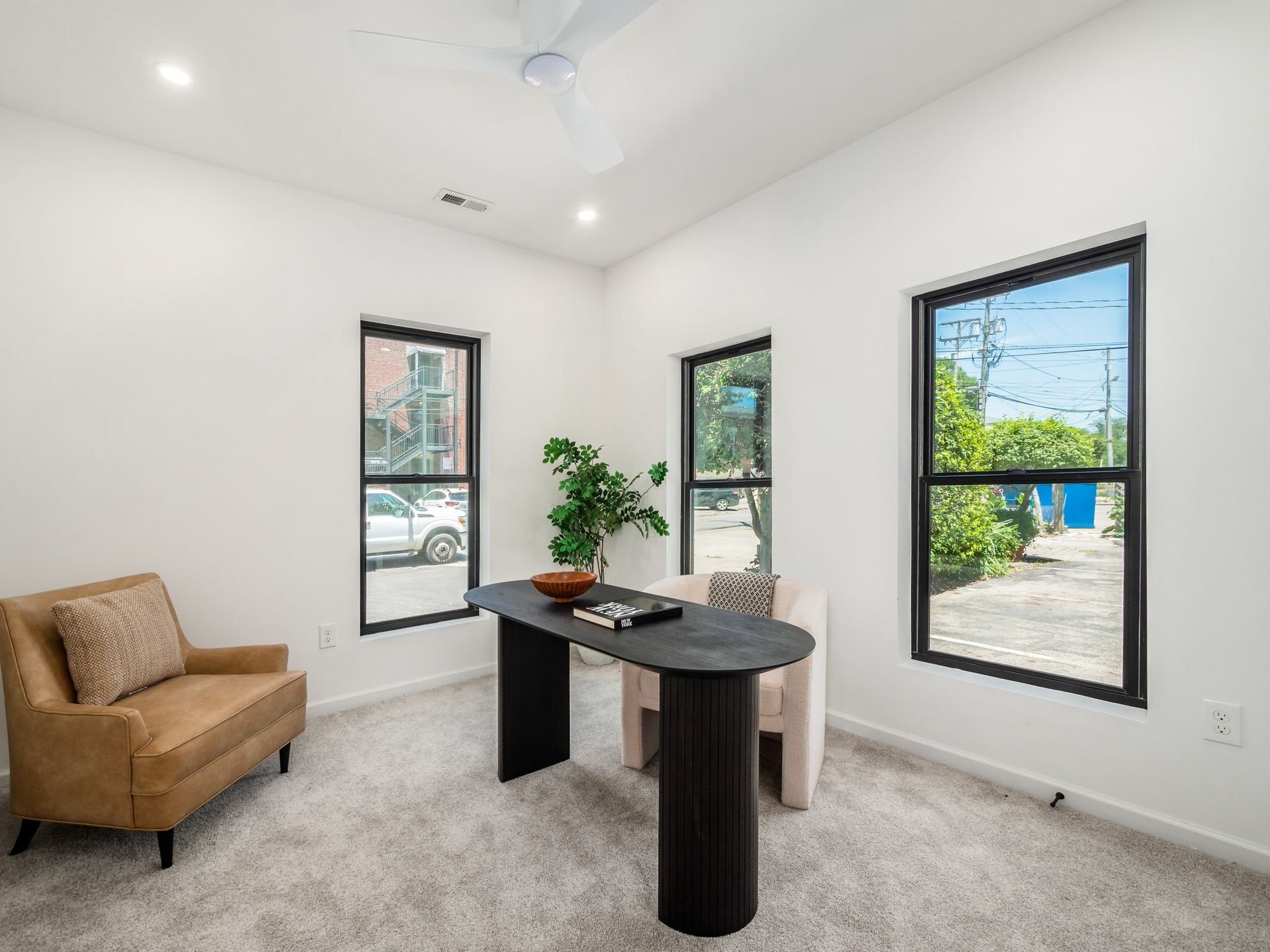 Minimalist office space with a small black desk, tan armchair, and beige chair, set against three windows with outdoor views. A potted plant and decorative bowl are on the desk, on a carpeted floor.