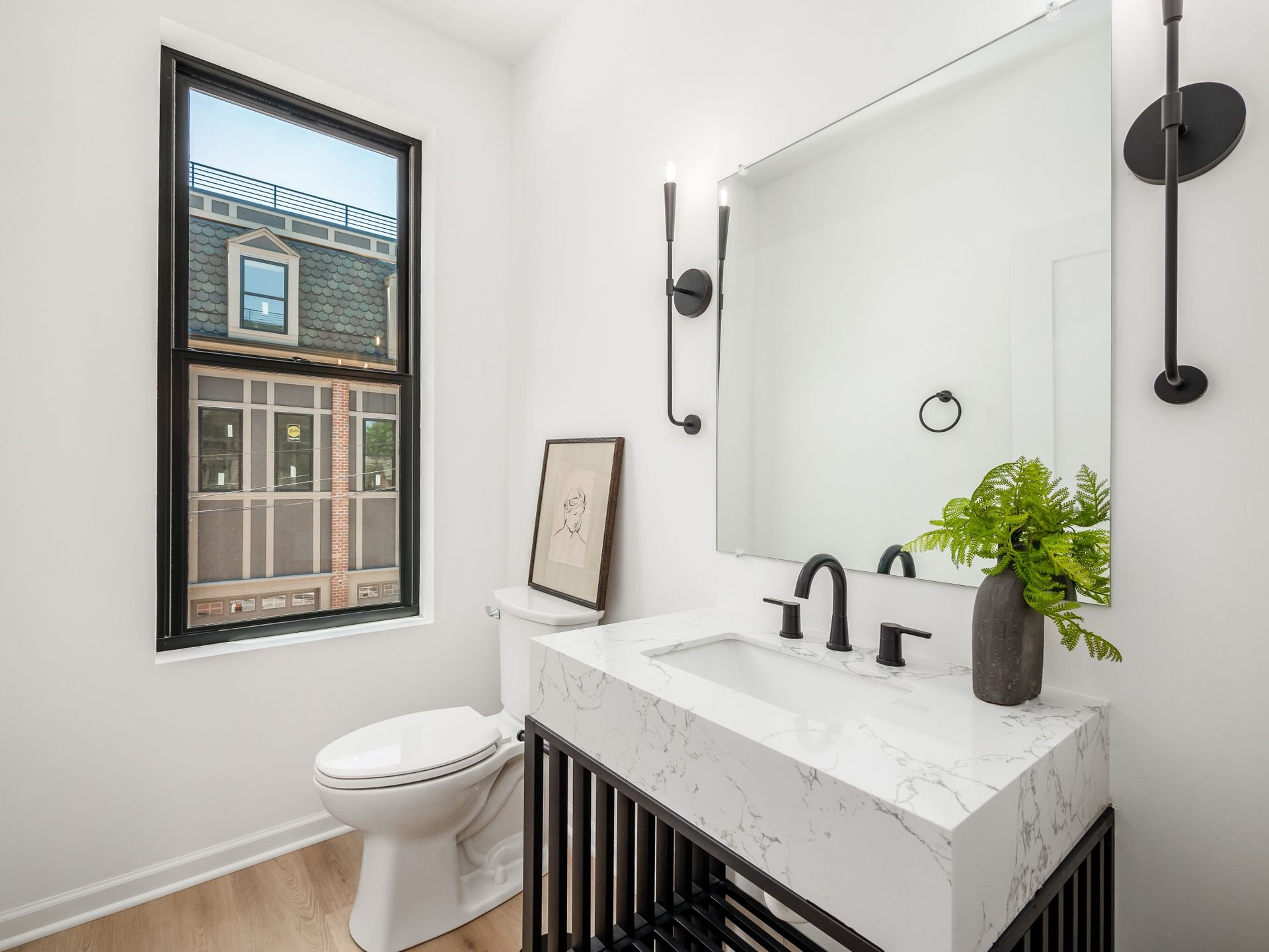 Modern bathroom with a white marble sink, black fixtures, a large mirror, a black wall-mounted light fixture, a window with a view of neighboring buildings, and a small framed sketch leaning against the toilet.