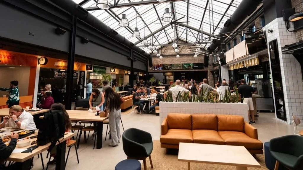 Interior of a busy food hall with people dining and socializing, featuring diverse seating options and a bright, industrial-style design with a glass roof.