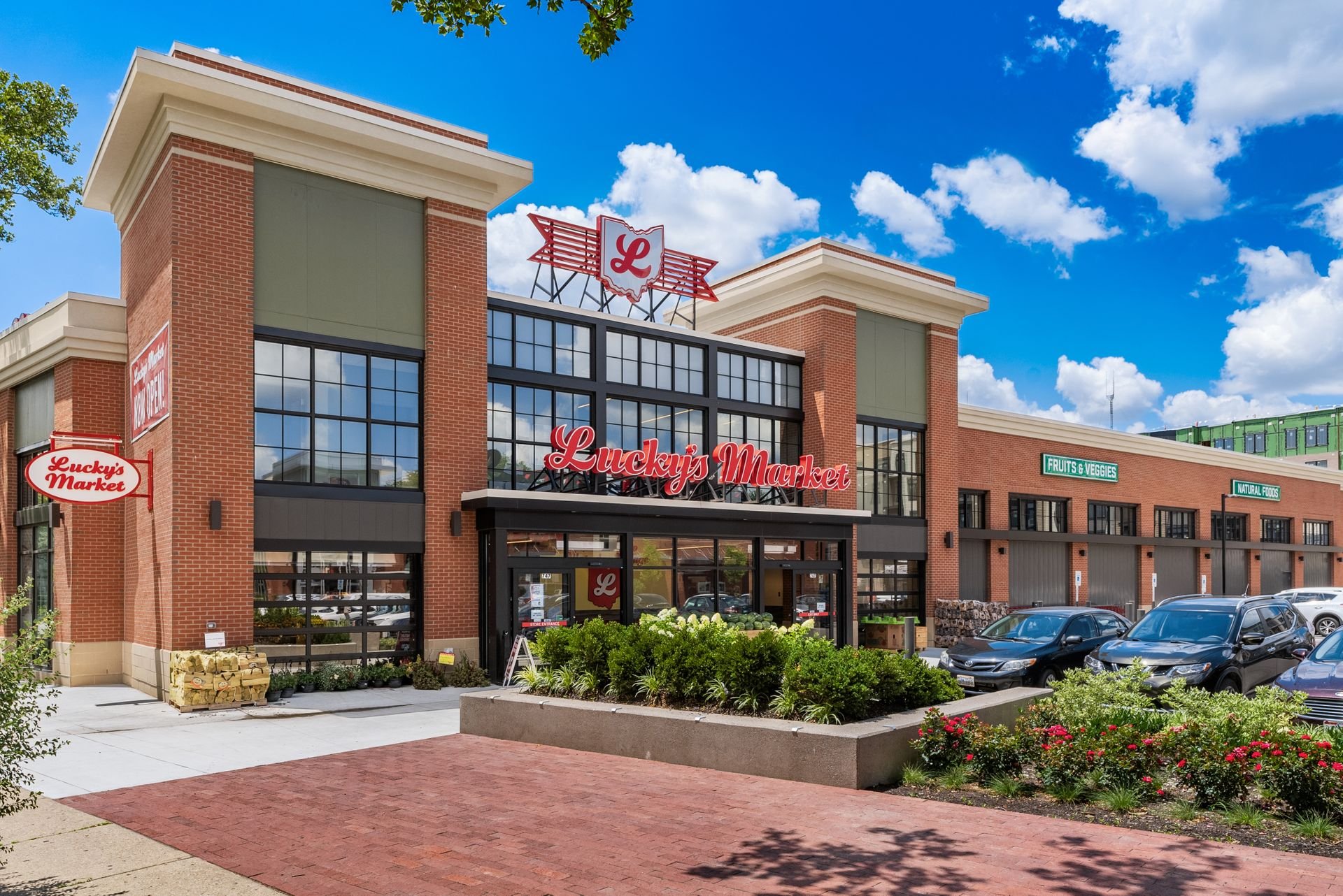 Exterior of Lucky's Market grocery store with brick facade, large windows, and red signage, surrounded by greenery and parked vehicles under a blue sky with clouds.