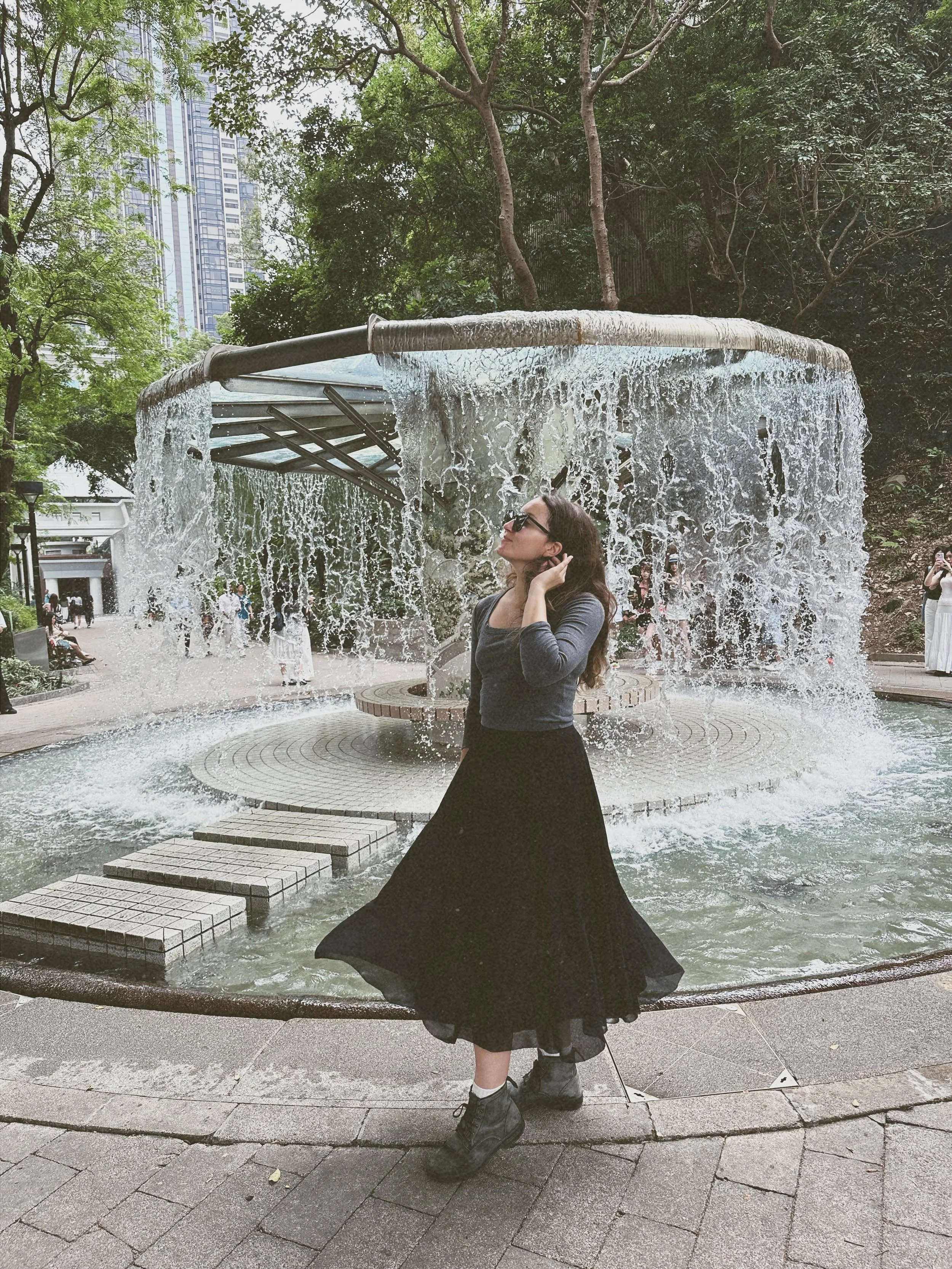 Amy Suto standing in front of the circular waterfall fountain at Hong Kong Zoological and Botanical Gardens, surrounded by lush greenery and city skyscrapers — a serene urban nature moment featured in her Hong Kong travel guide on AmySuto.com.