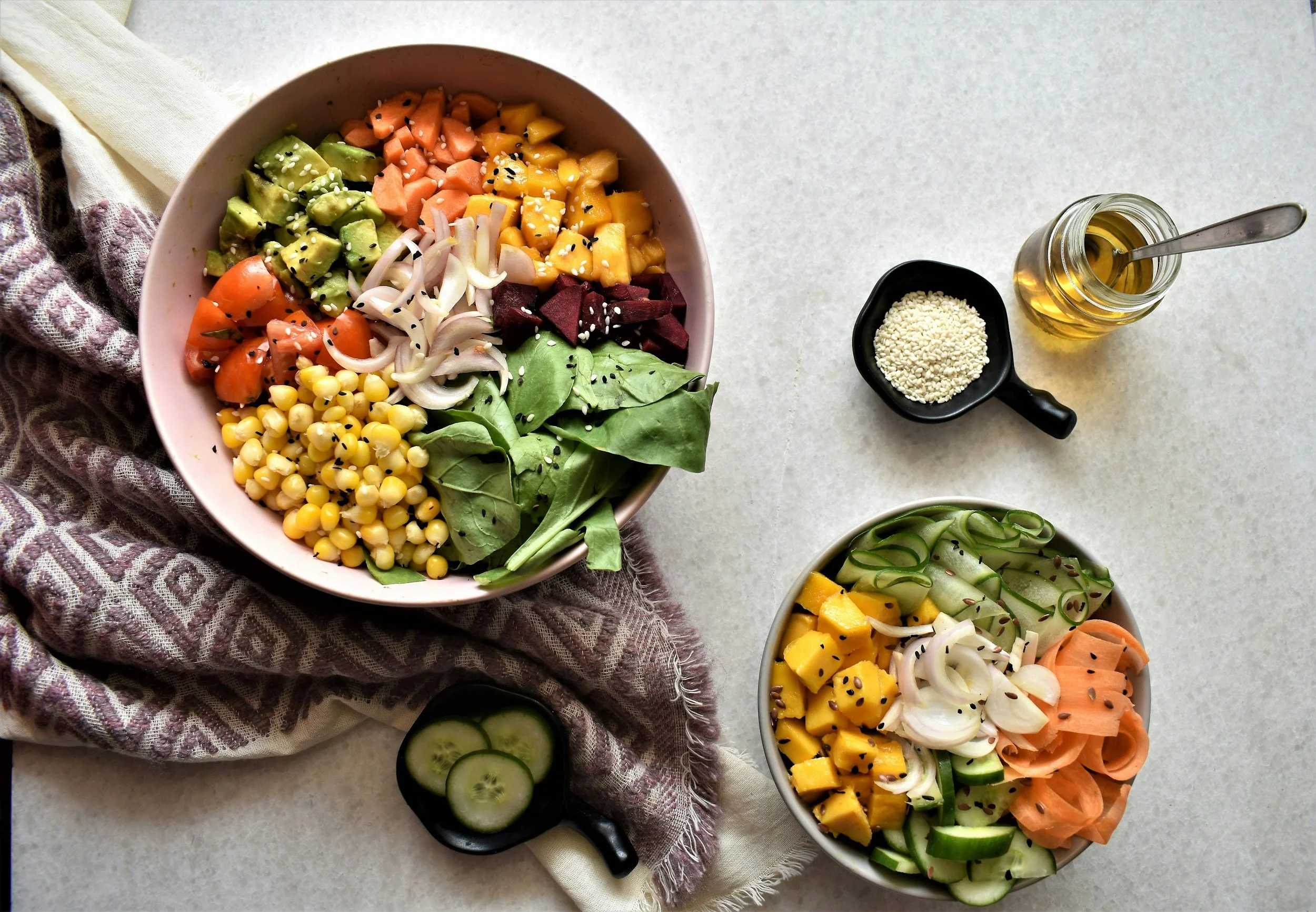 Two bowls of fresh salad with sliced cucumbers, mango, avocado, cherry tomatoes, onion, and beets, sprinkled with sesame and black seeds. A small jar of vinaigrette and a small cup of sesame seeds are on the table.