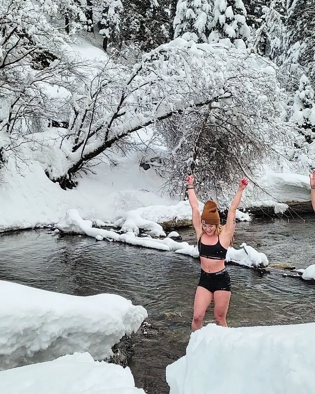 A woman in black sportswear and a brown beanie hat standing in a snowy river with arms raised in celebration, surrounded by snow-covered trees and rocks.