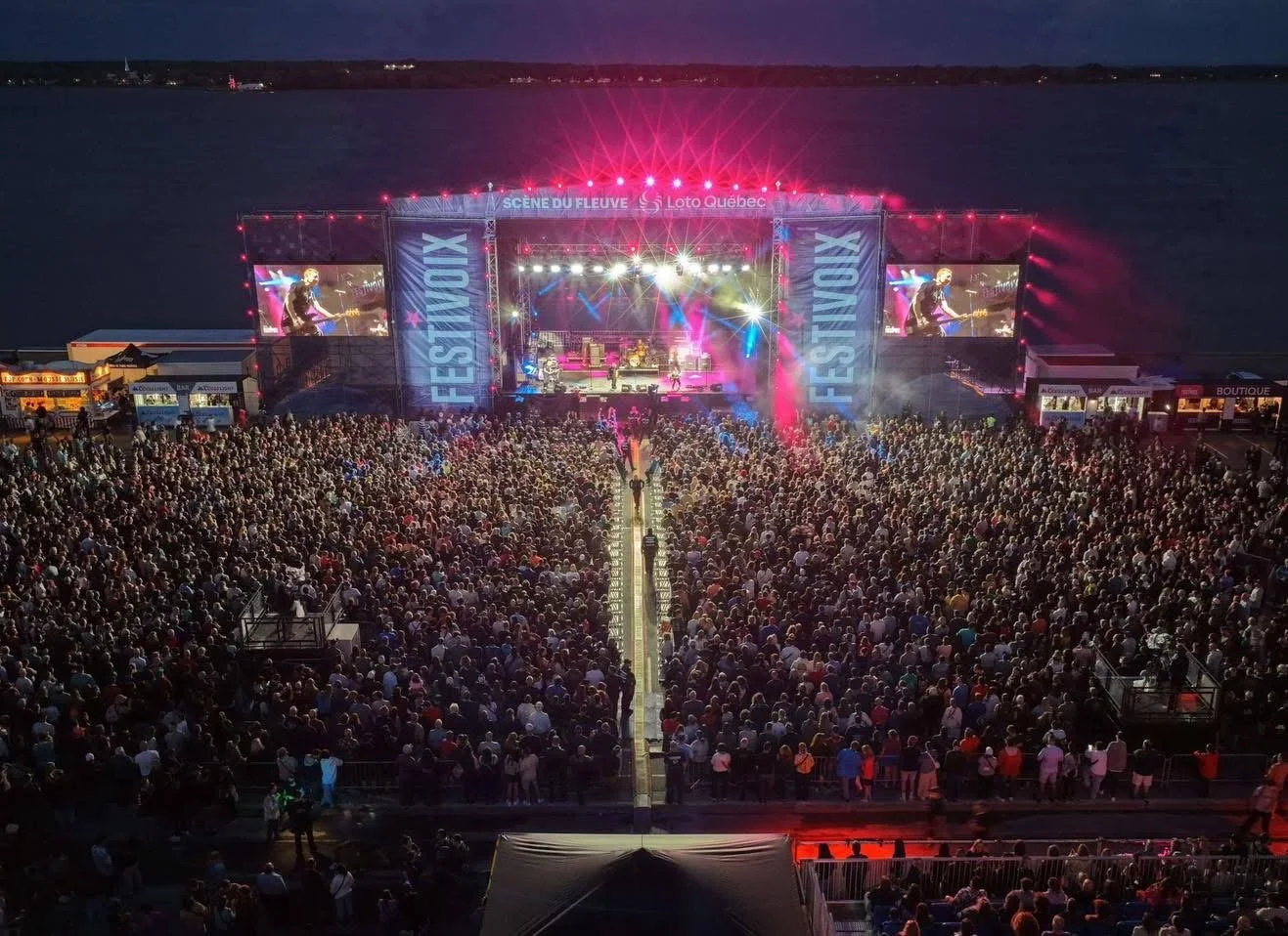 An outdoor music festival by a river at night with a large crowd watching a live concert on a bright, colorful stage. Large screens display the performer, and festival banners read 'Festival' and 'Scene du Fleuve'.