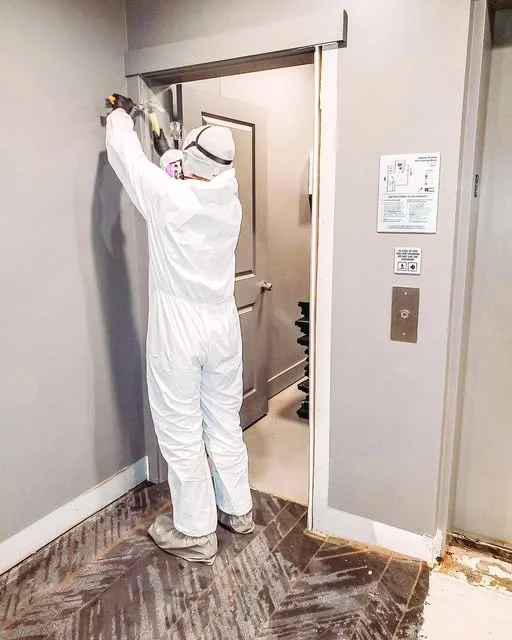 A hazardous materials cleaner wearing a full white protective suit, gloves, and a face shield sprays disinfectant on a wall next to an elevator door.